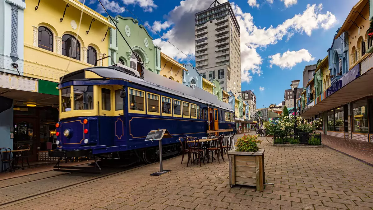 Christchurch Blue Tram on a brick road with colourful buildings behind, benches and plants in the foreground with a blue sky  Main Image