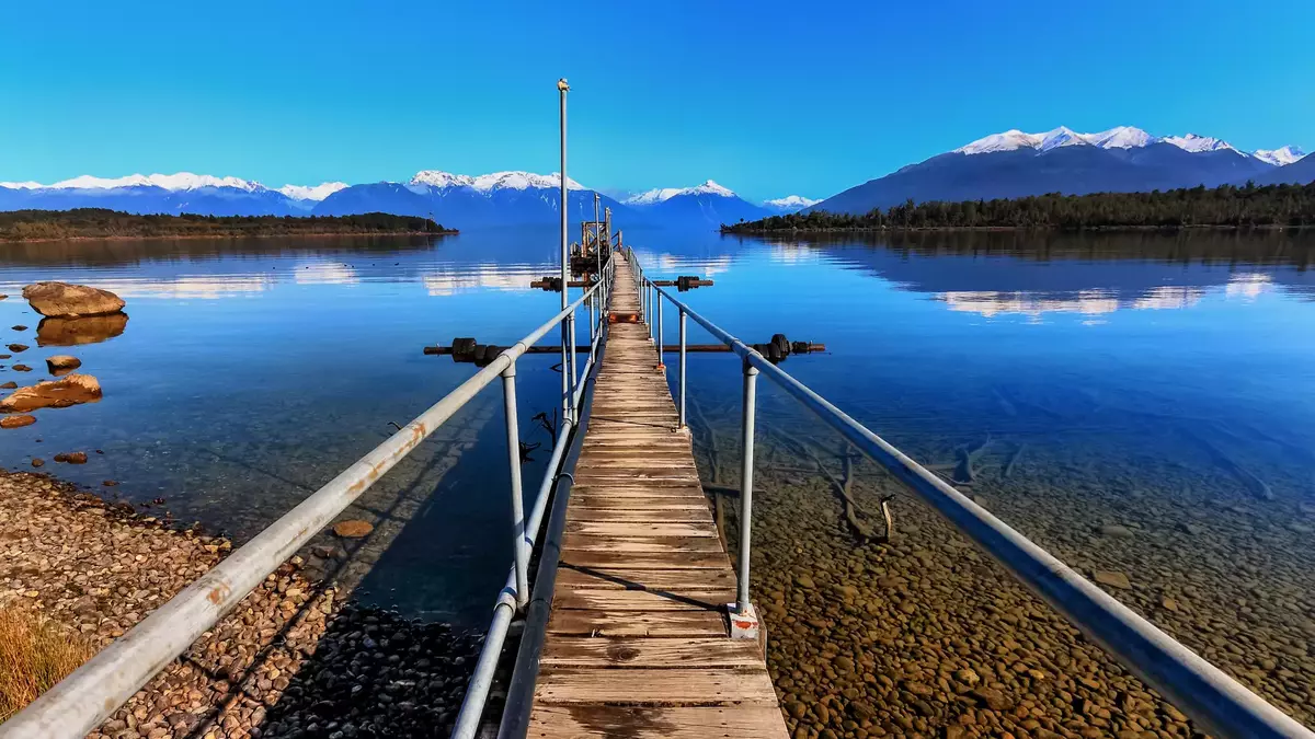 Wooden jetty stretching into a lake with clear water and snowcapped mountains under a bright blue sky. Main Image