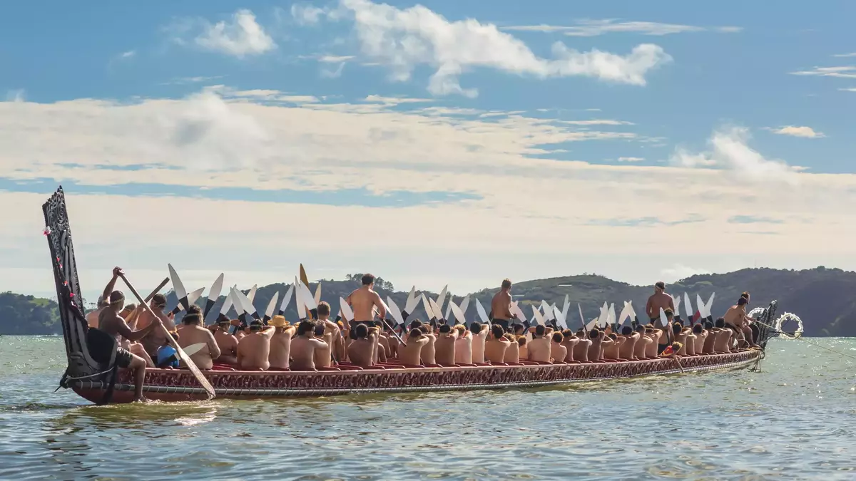 People paddling a traditional waka canoe on water with hills and blue sky in background. Main Image