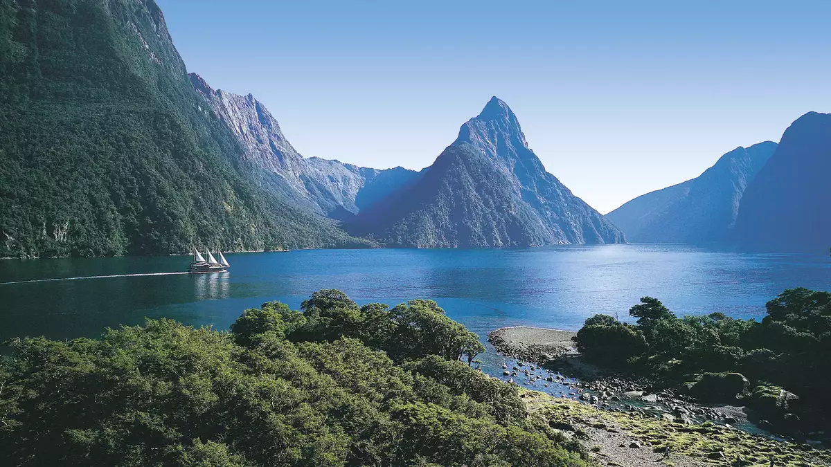 Sailboat on calm blue water surrounded by towering green mountains under clear sky. Main Image