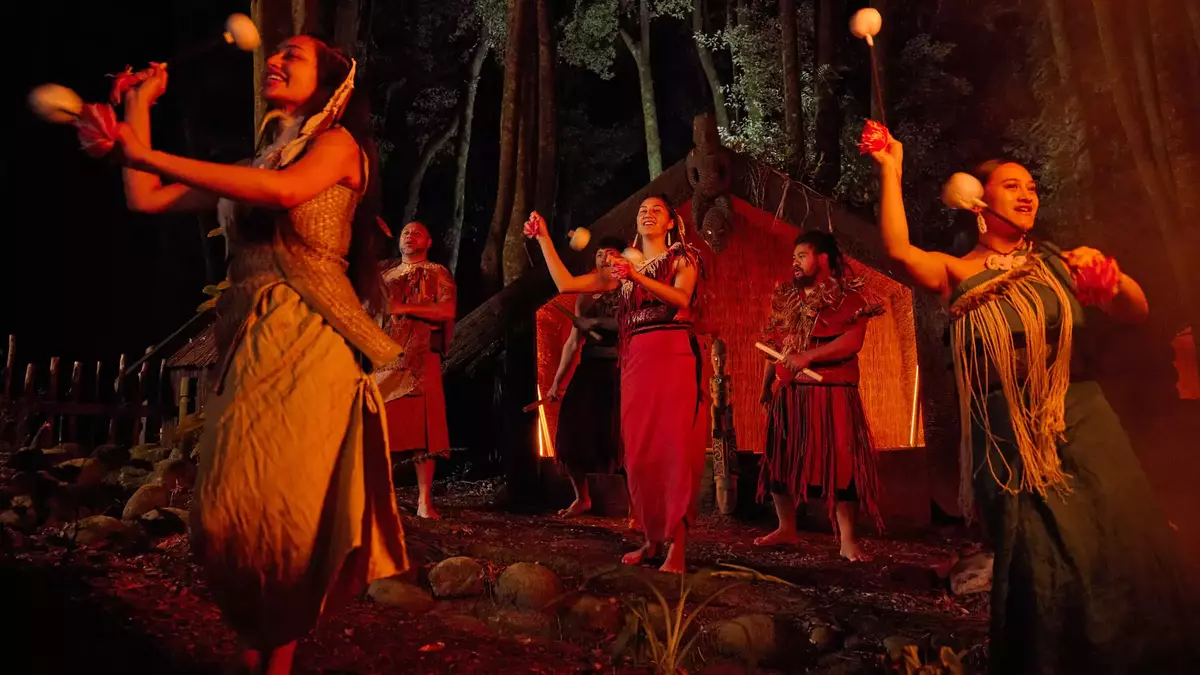 Māori performers in traditional dress perform a poi dance on a forest stage at night, with a carved wharenui and towering trees illuminated in the background. Main Image