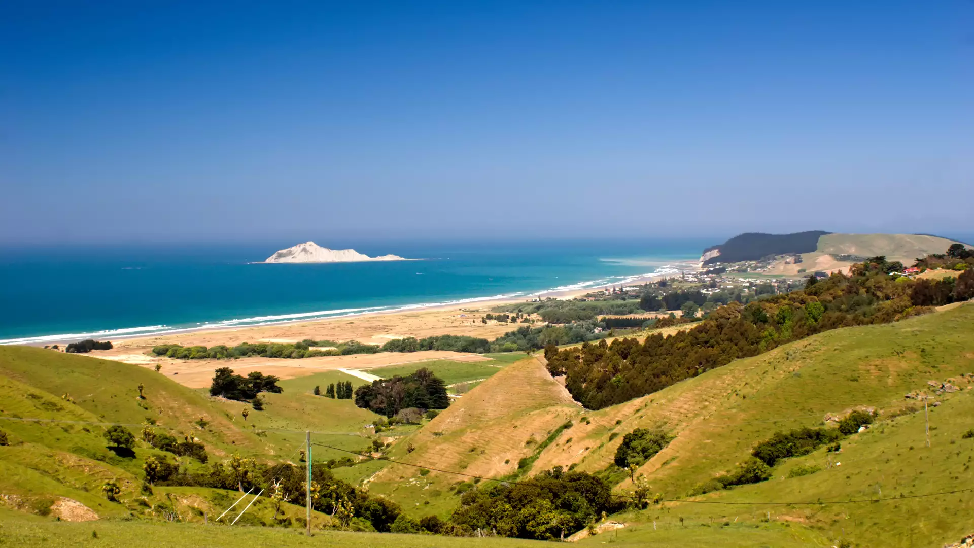 View of green hills, sandy beach, ocean, and a small island off the coast under a clear blue sky. Main Image