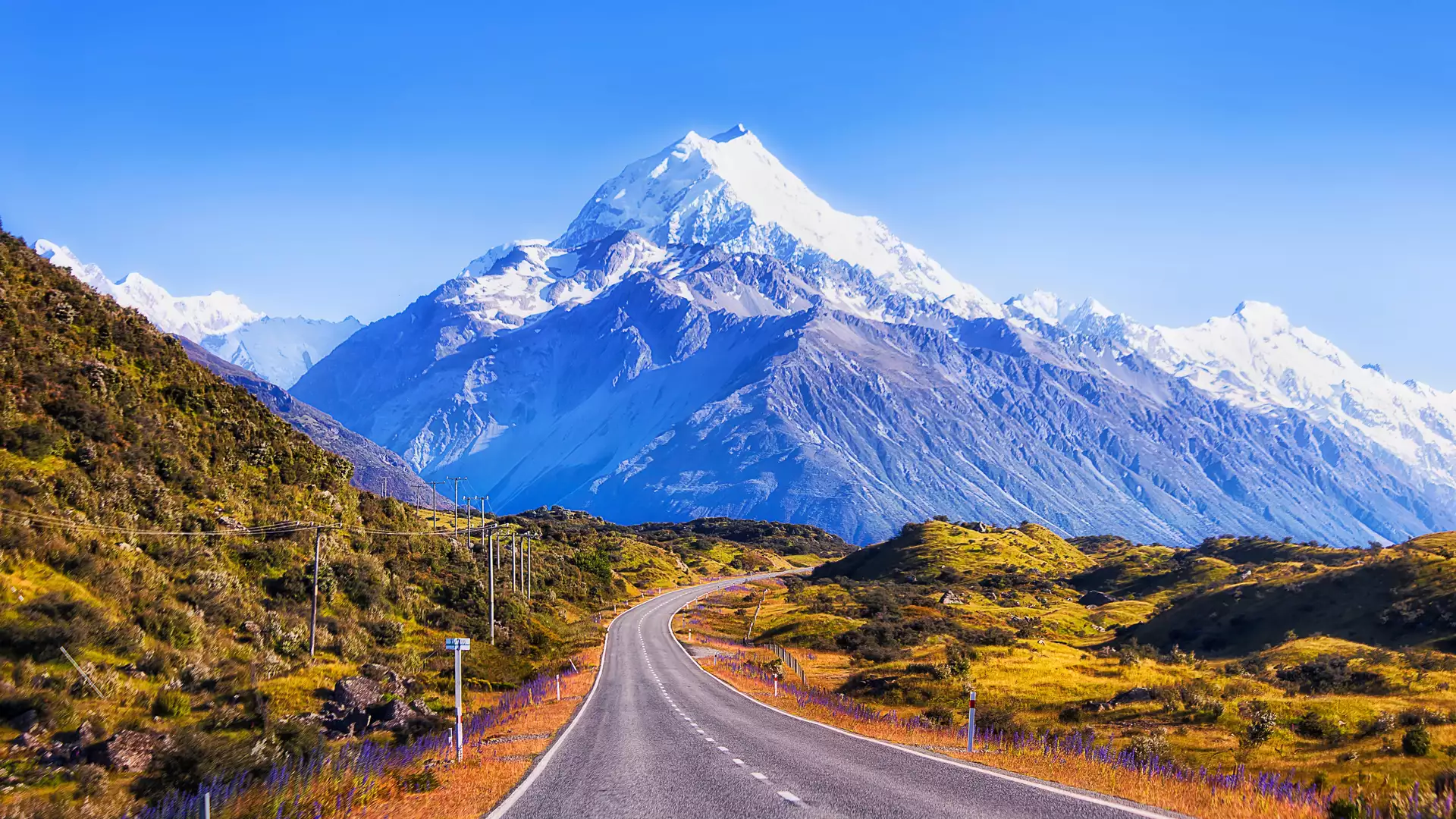 A Long curing road leading up to Mt Cook with snow on the top and green verges Credit: Mt Cook Road Main Image