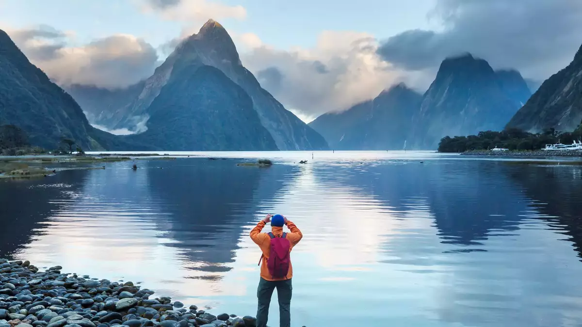 A person in an orange coat looking out over Milford Sound with water and hills in the background Main Image