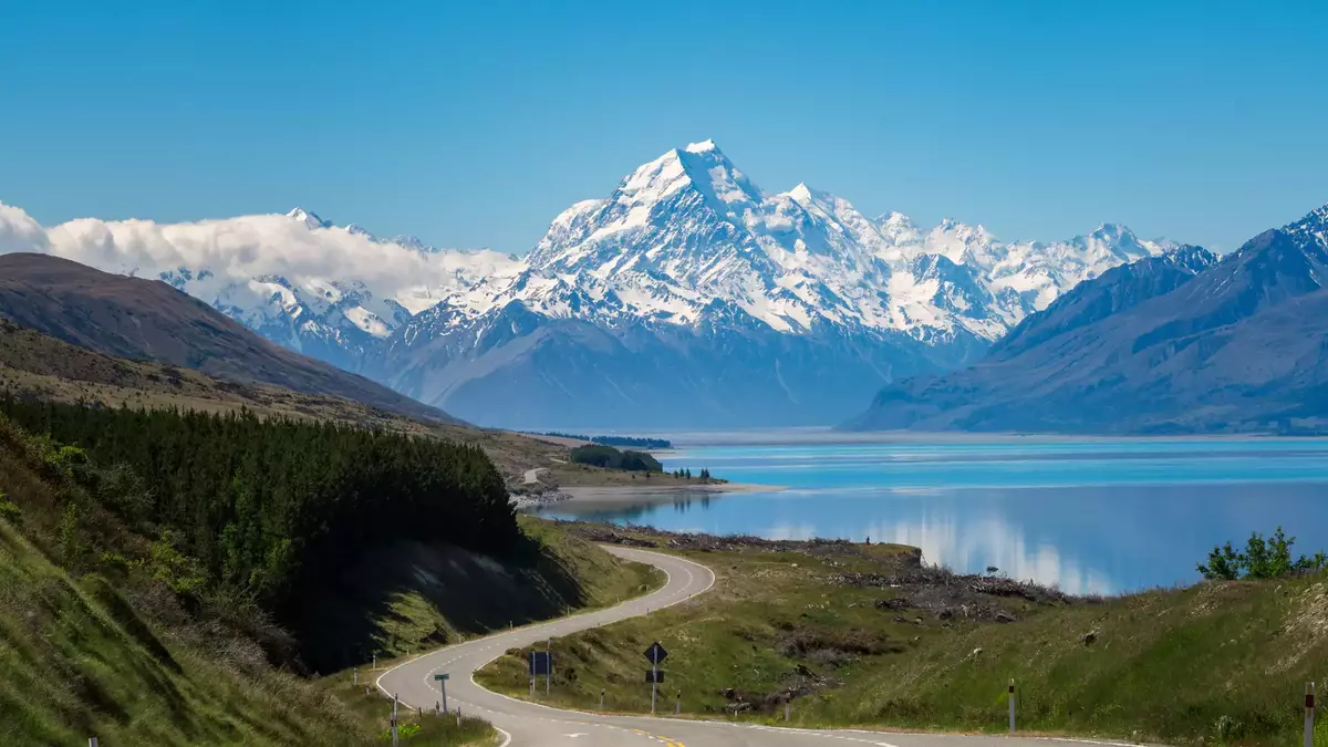Curved road leading towards snow-covered Aoraki/Mt. Cook mountains by a reflective lake. Main Image