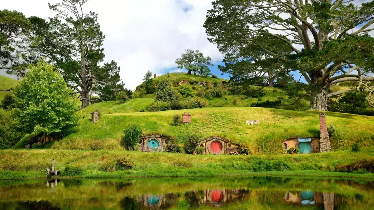 Colorful hobbit house doors built into green hills with large trees and a pond reflecting the scene Main Image