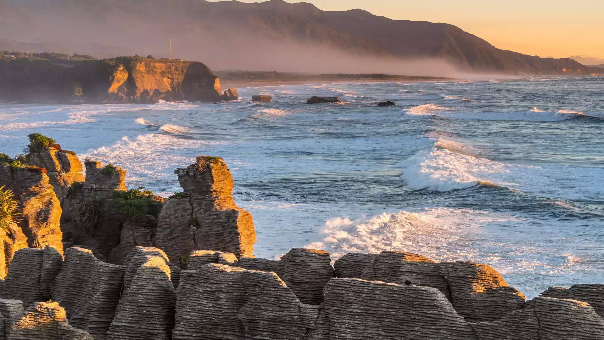 Pancake Rocks coastal rock formations at sunset with waves and mountains in the background Credit: New Zealand Fine Touring Main Image