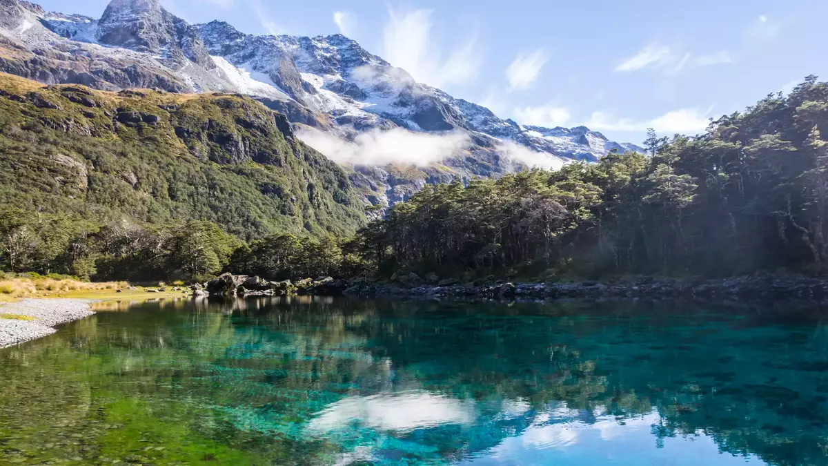 Clear lake with forest and snow-capped mountain reflections under blue sky Main Image