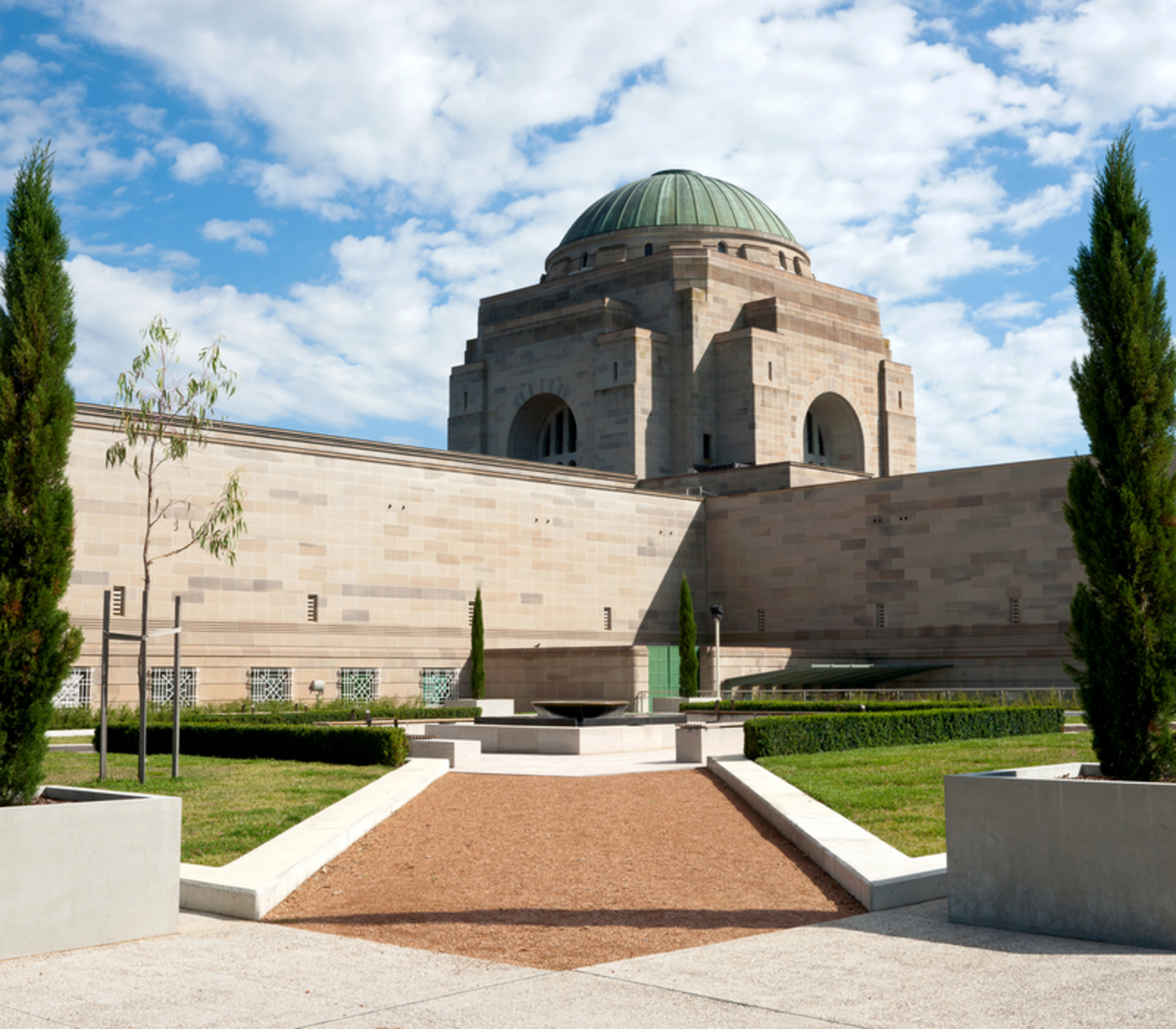 Exterior view of the Australian War Memorial in Canberra with manicured gardens and cypress trees under a bright sky.