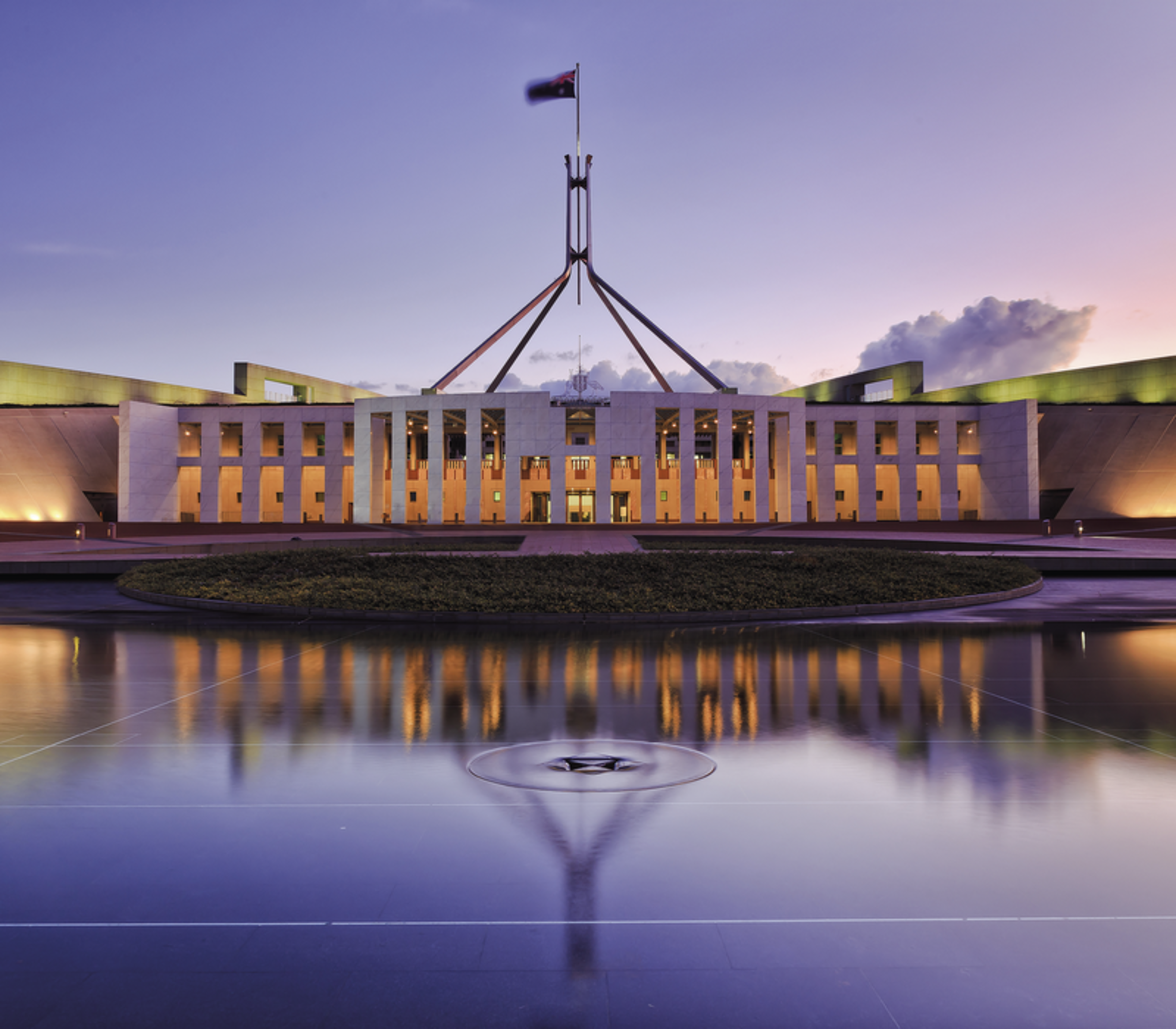 The facade of Parliament House in Canberra reflected in a still pool during a purple and orange sunset.