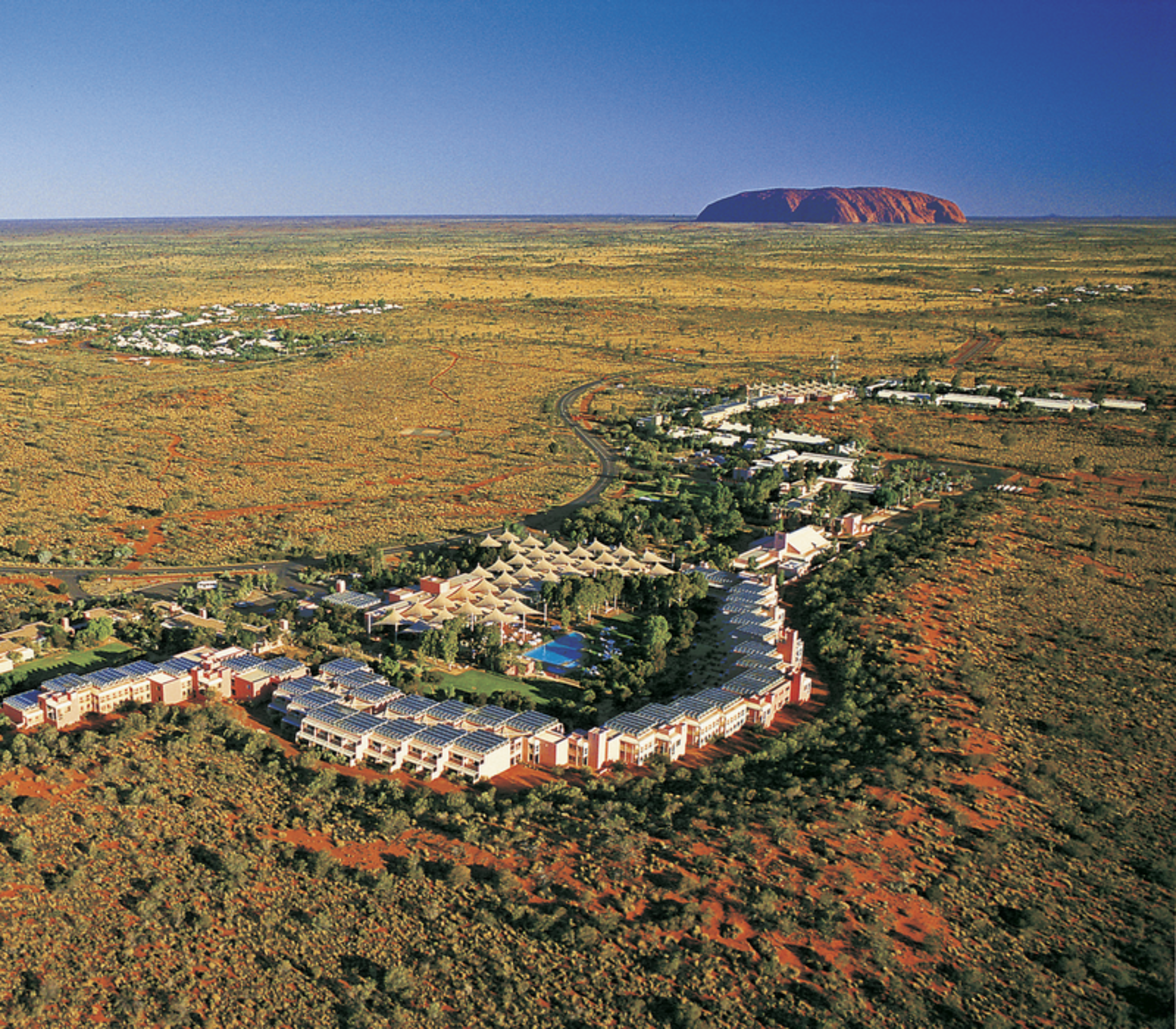 High-angle aerial view of the desert resort buildings and swimming pools with Uluru visible in the far distance under a clear blue sky.