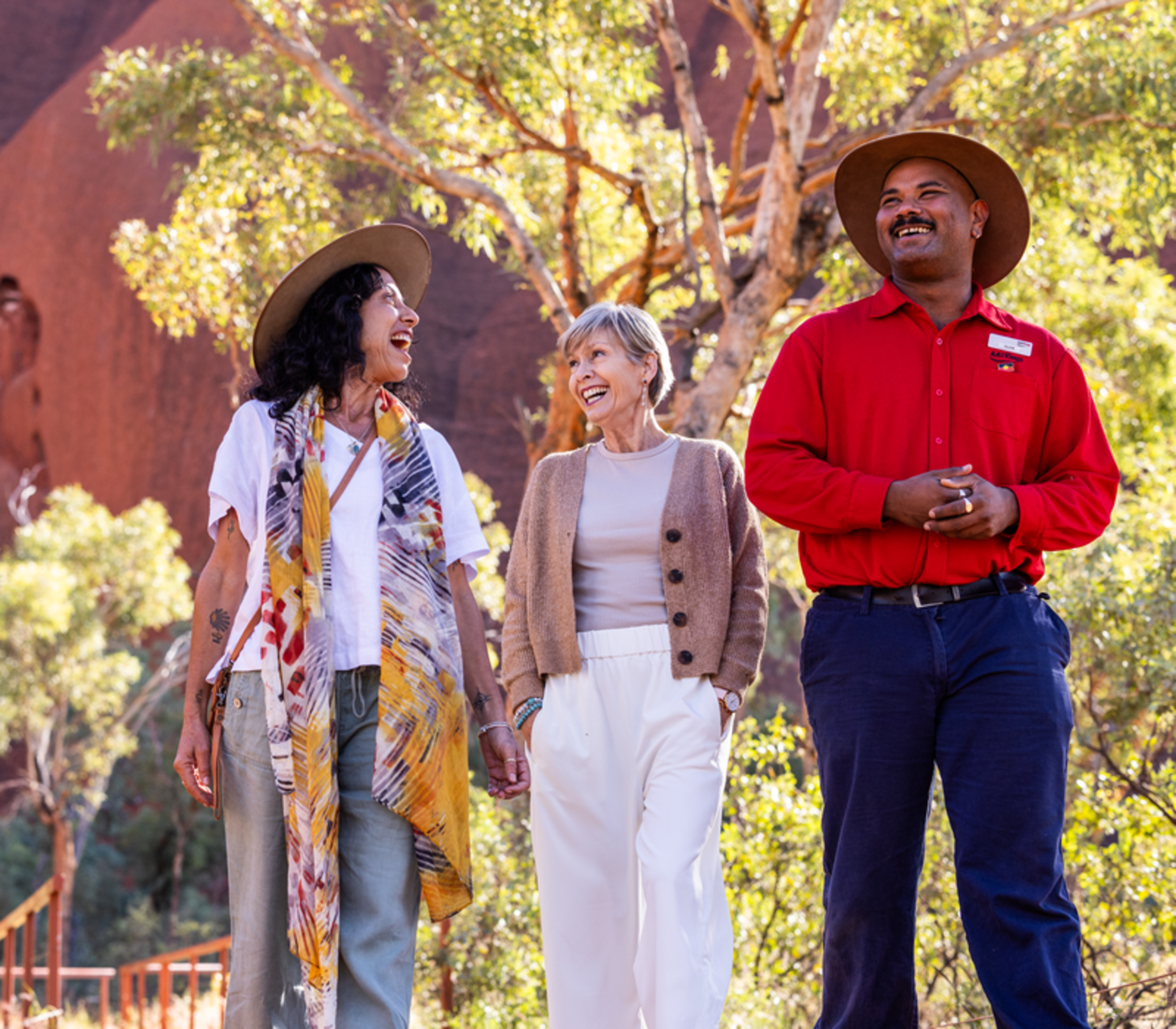 A guide and two female travellers laughing while walking past a native gum tree at the base of Uluru.