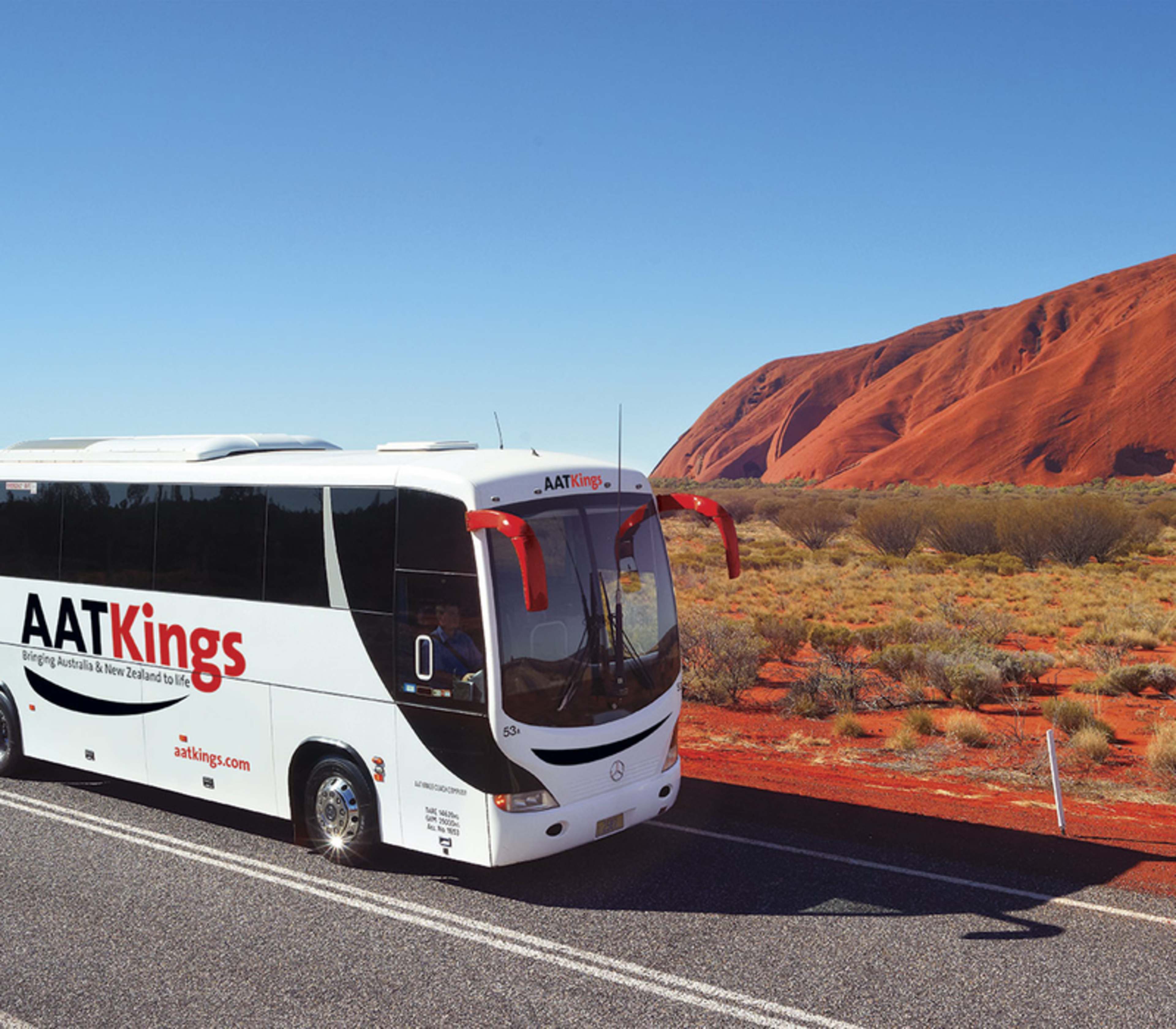 A white AAT Kings tour bus driving on a paved road through the red desert with Uluru looming on the horizon.
