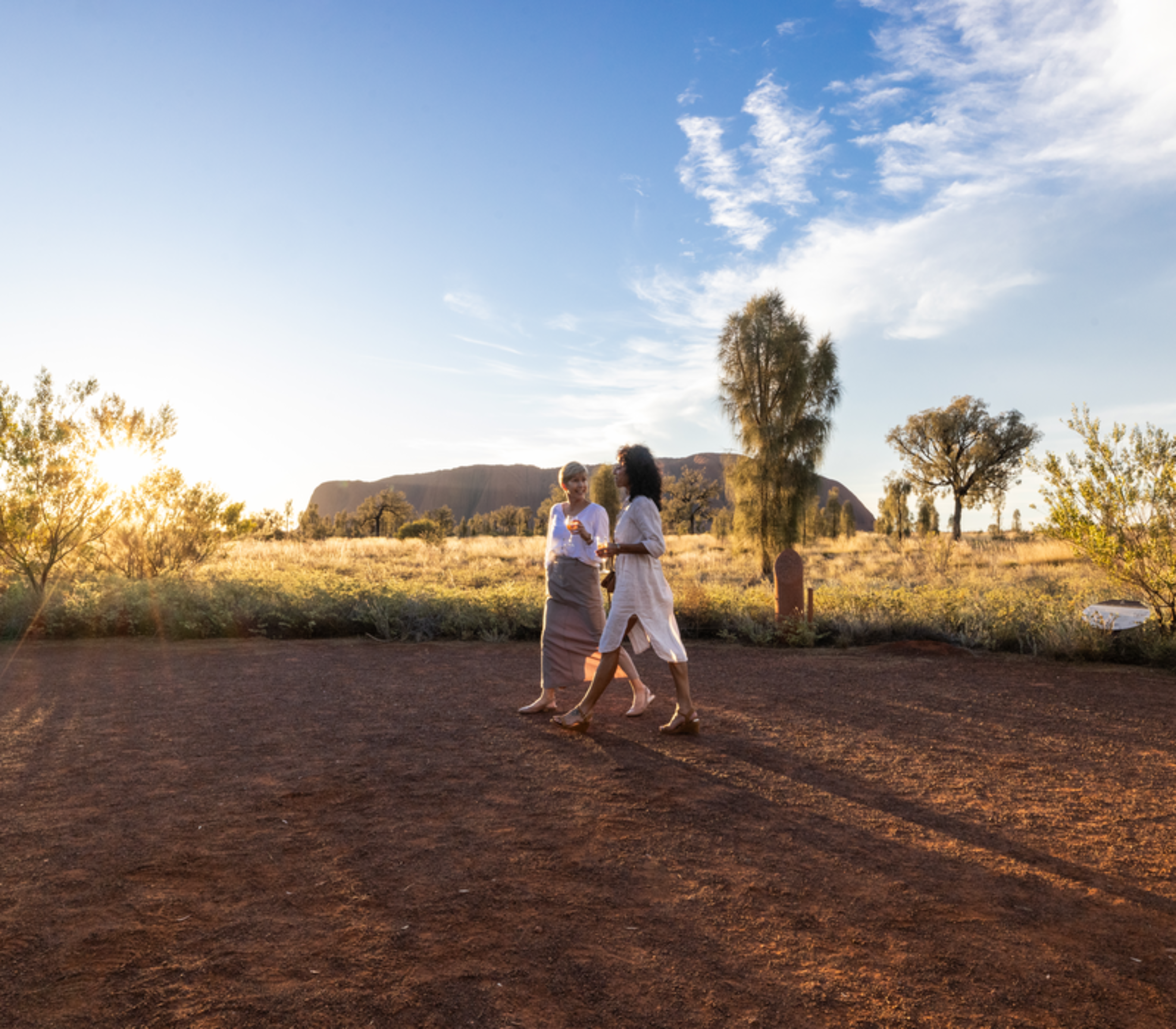 Two female travellers walking on a red dirt path in the desert with Uluru in the far distance.