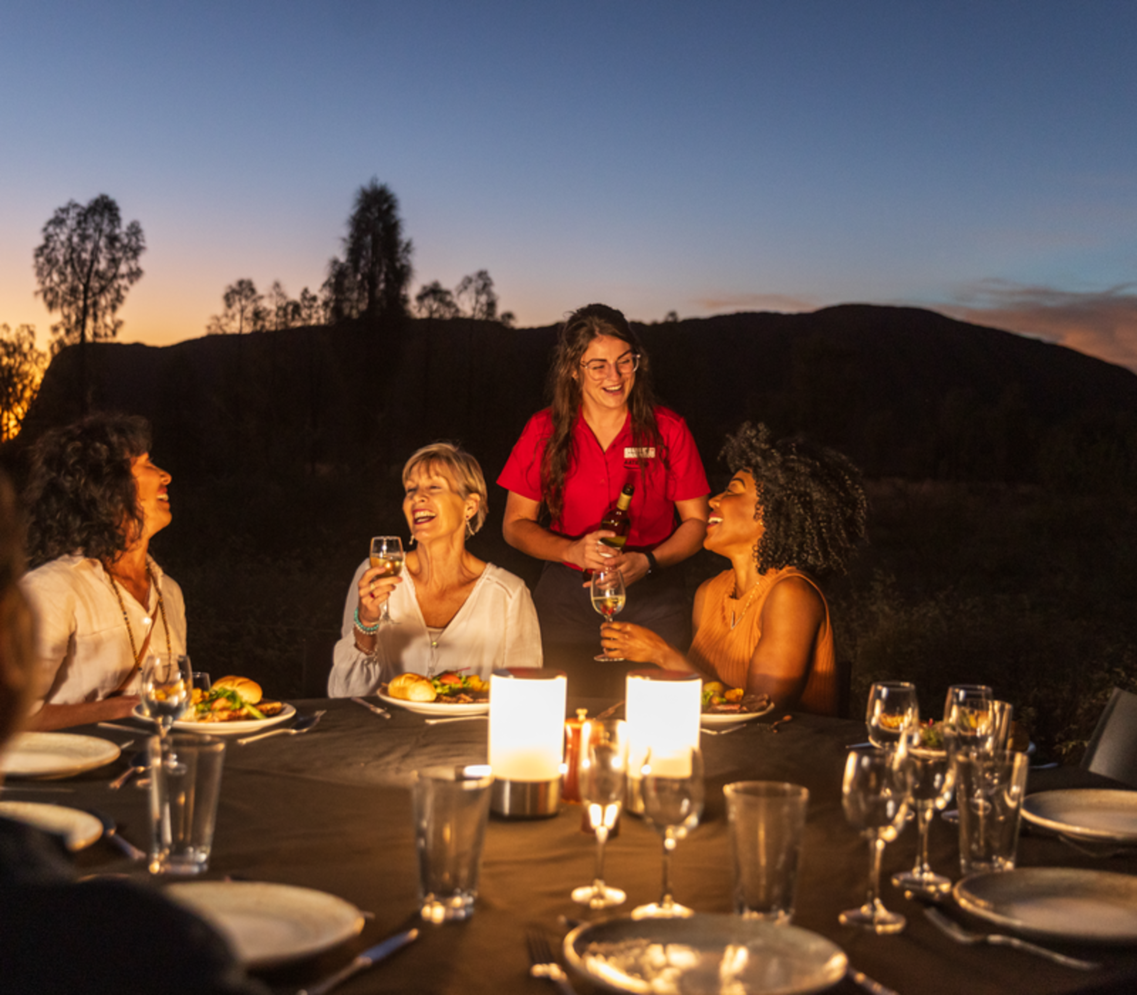 Travellers enjoying a candlelit dinner and wine at an outdoor table in the Red Centre at dusk.