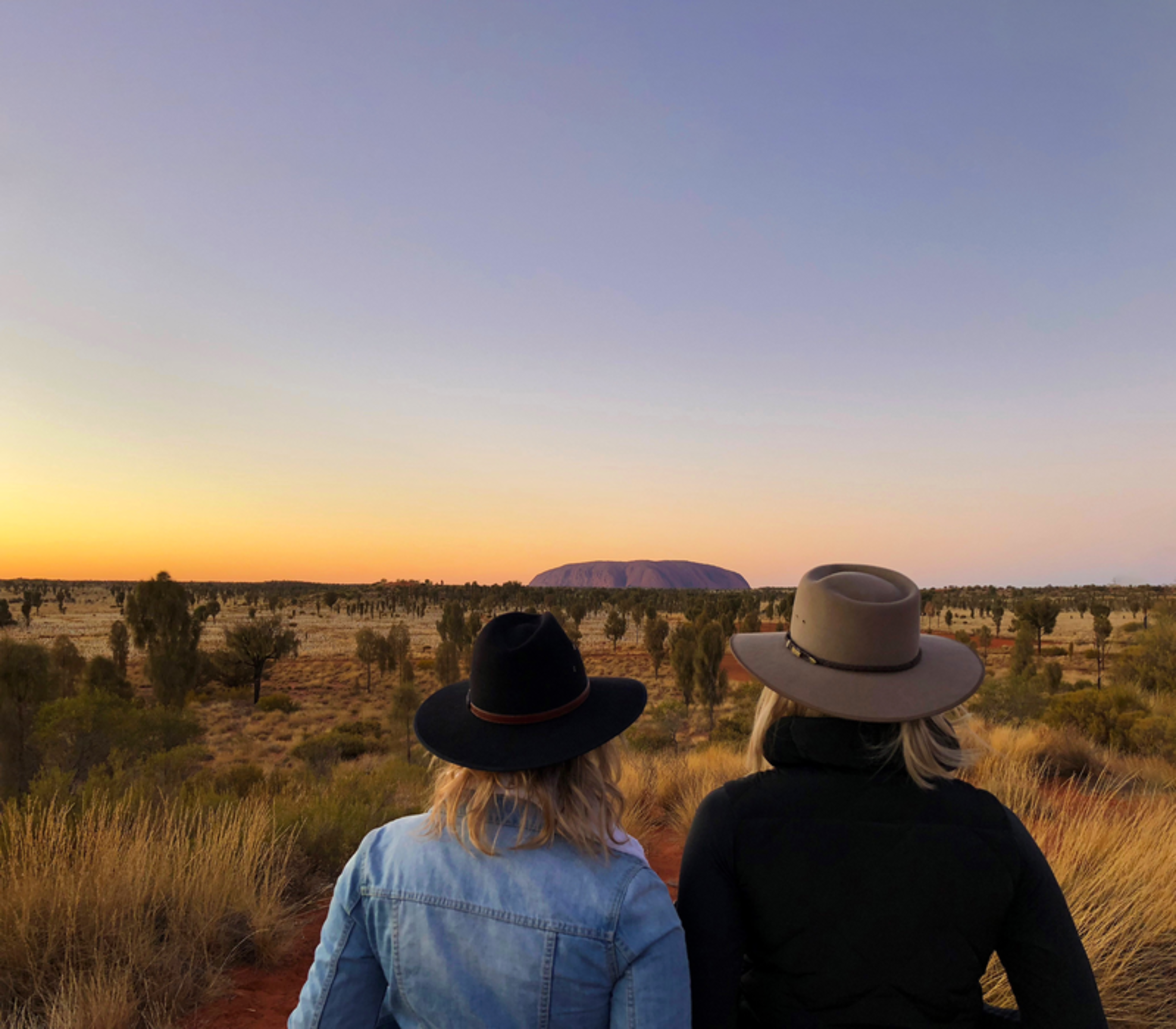 Back view of two women in hats looking out across the desert at Uluru during a vibrant golden sunset.