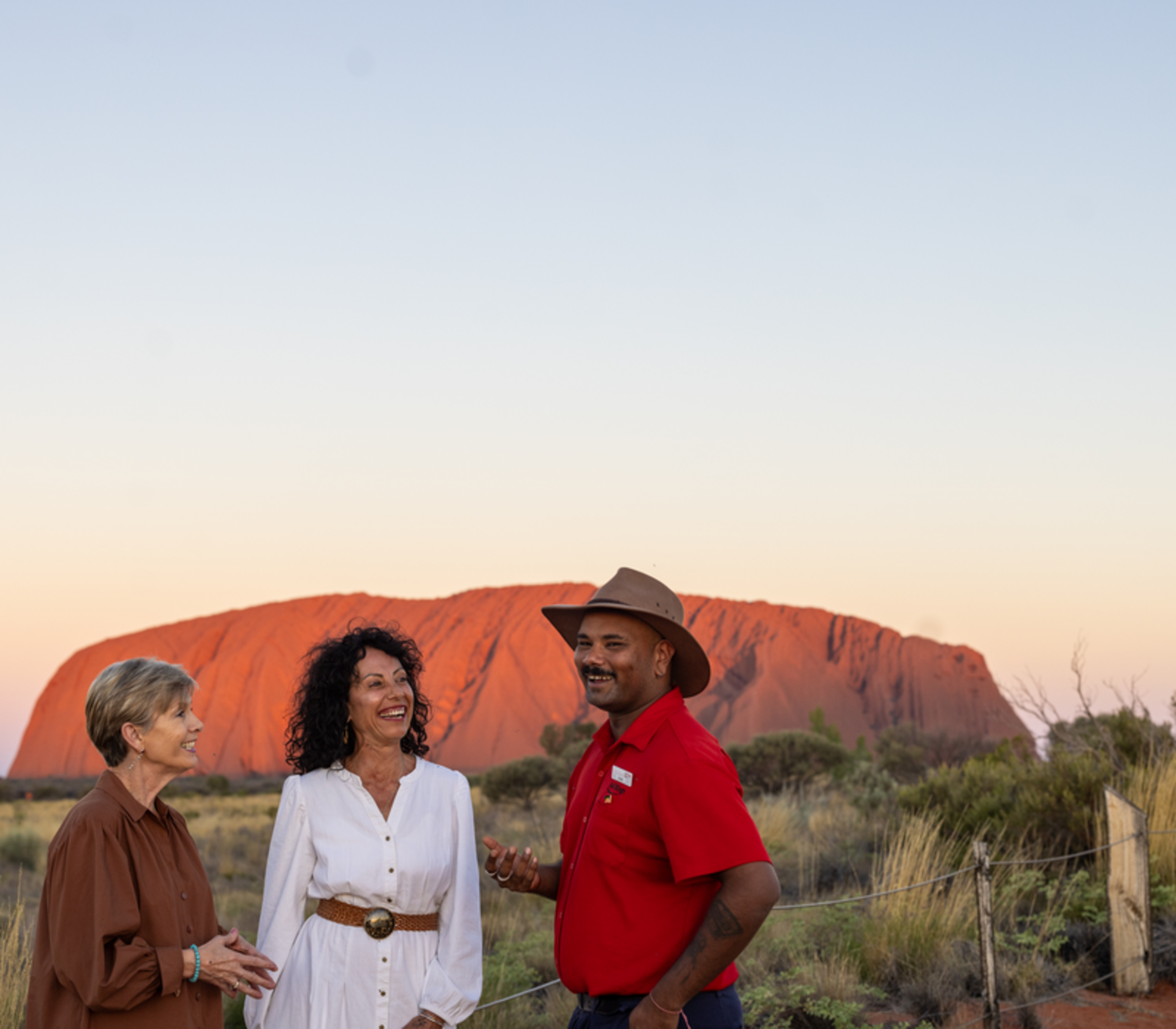 A guide talking to two women with the vibrant red rock of Uluru in the background at sunset.