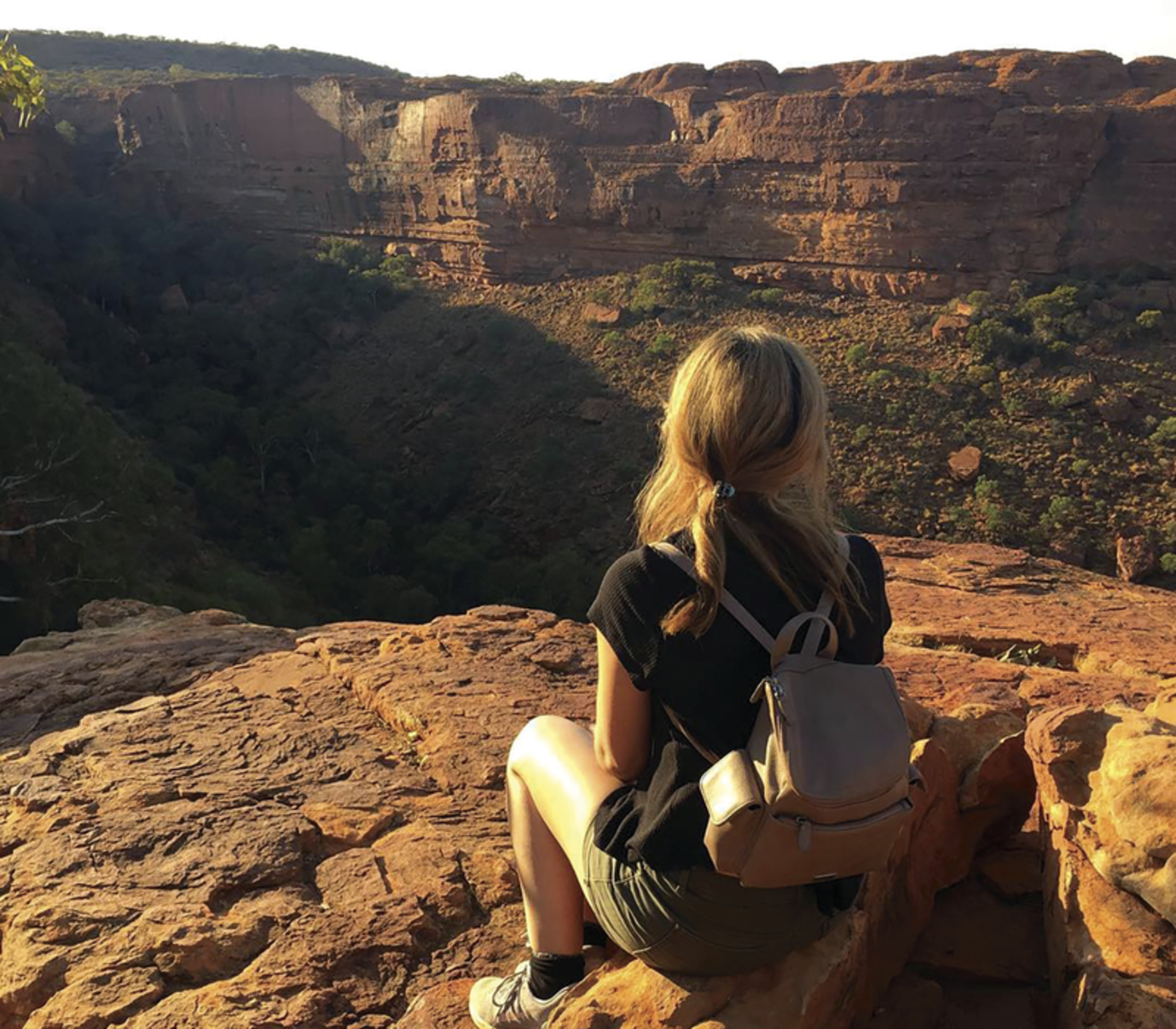 A person with a backpack sitting on a rocky cliff overlooking the deep red sandstone walls of Kings Canyon, Northern Territory.