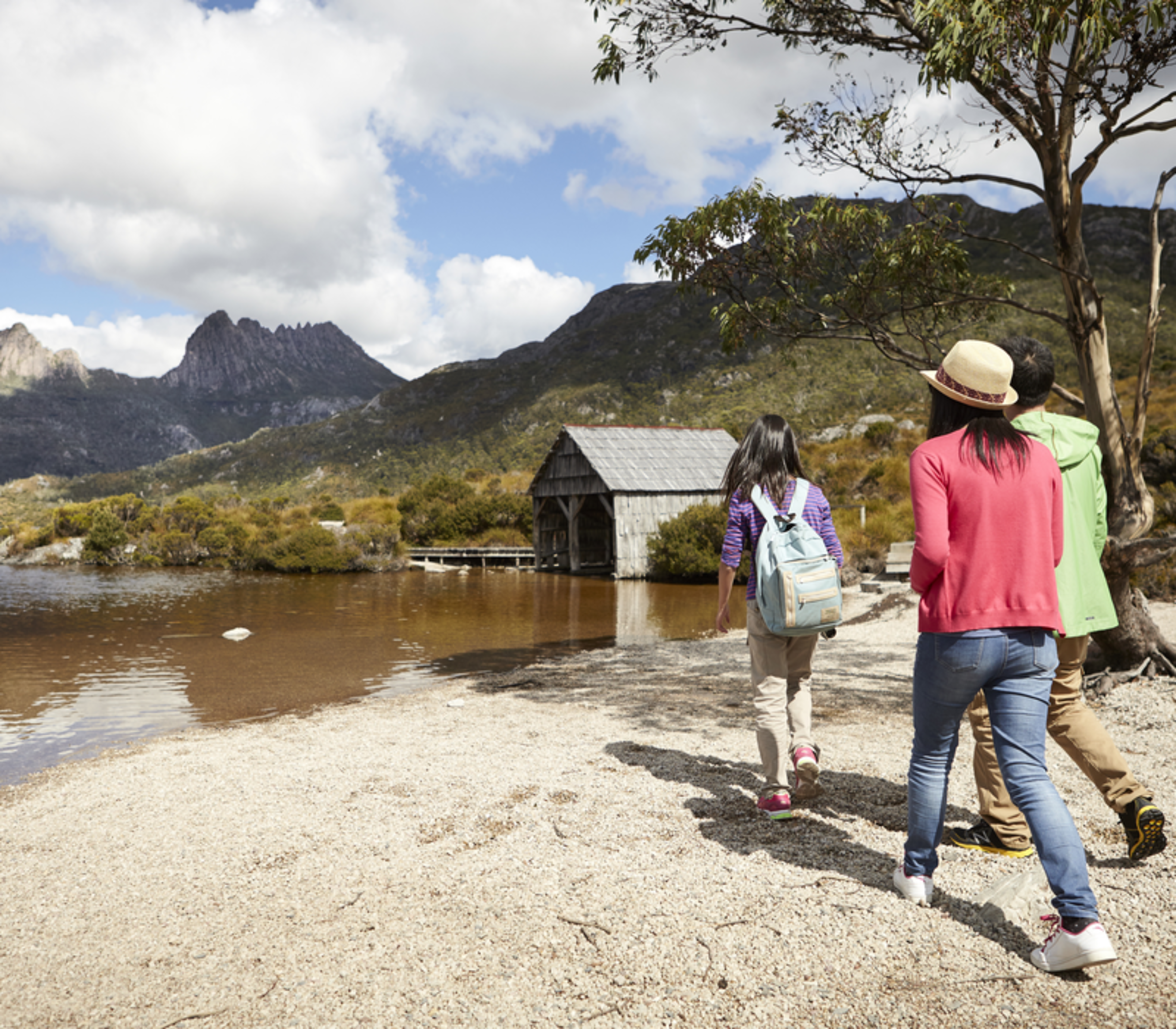 A group of tourists of three walking along the pebbled shore of Dove Lake towards the iconic boat shed at Cradle Mountain, Tasmania.