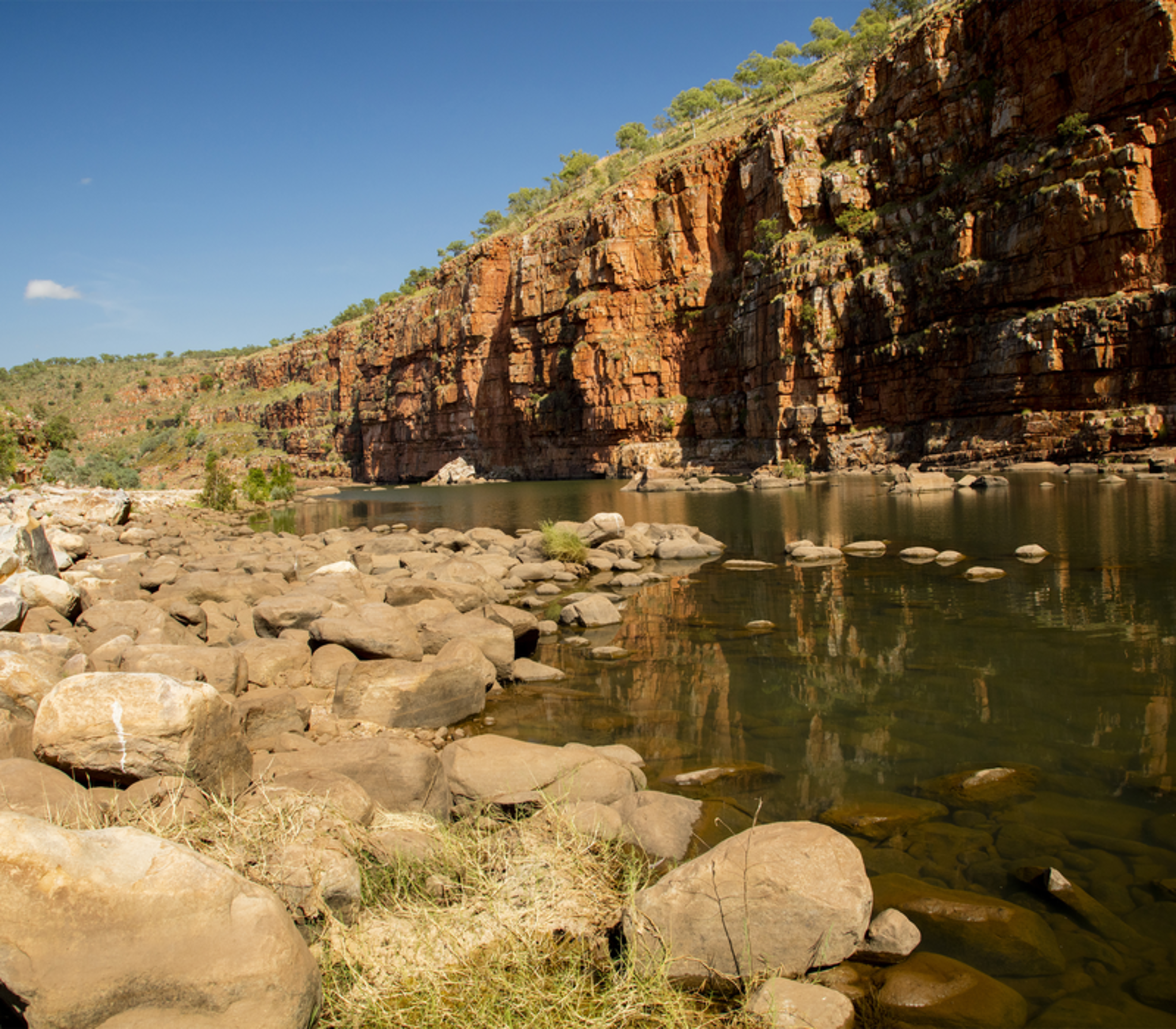 High red rock walls of a gorge reflecting in a river with boulders in the foreground, The Kimberley, WA.