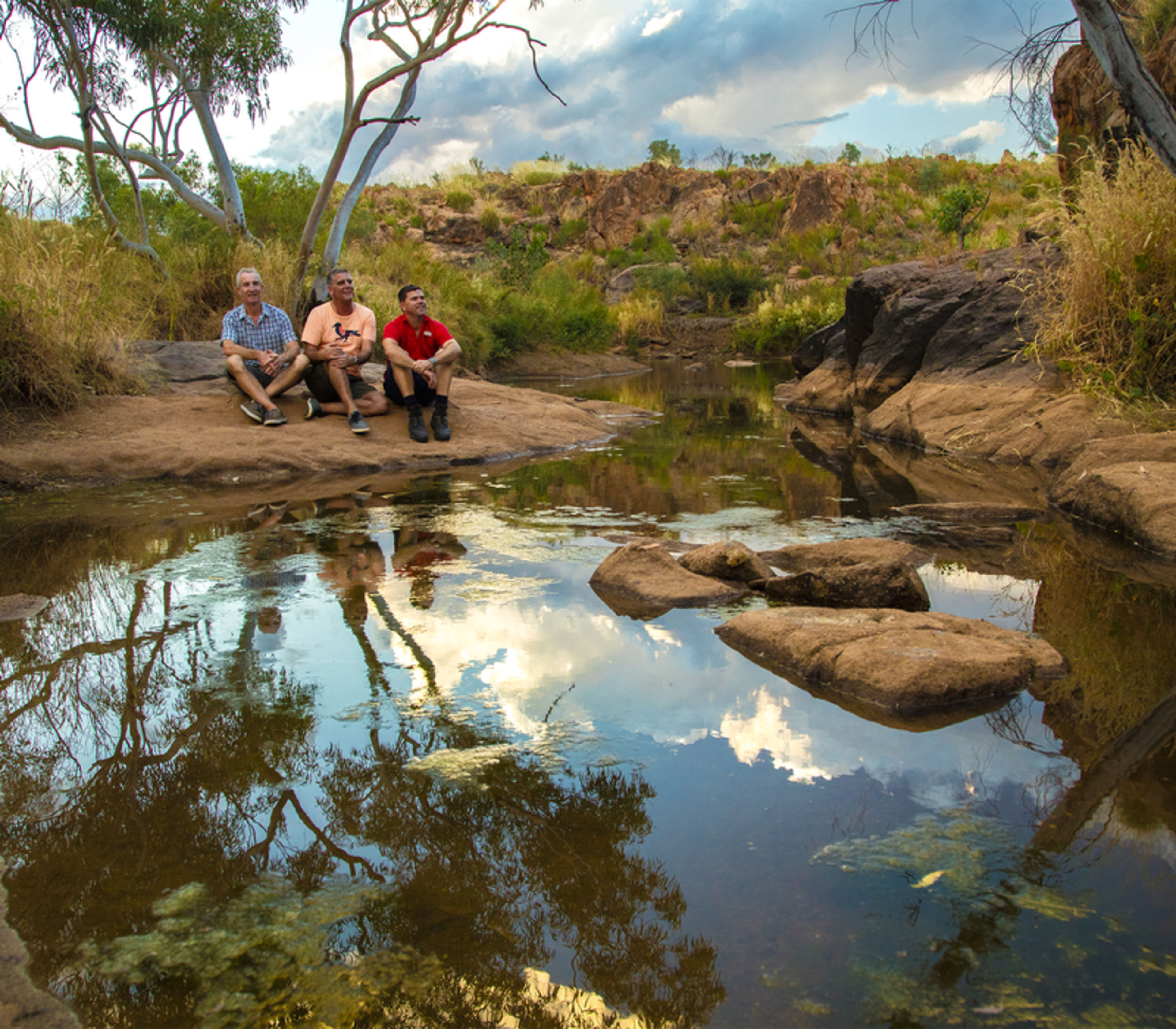 Three people sitting on a rocky bank next to a still pool of water in the Kimberley outback near Halls Creek at sunset.