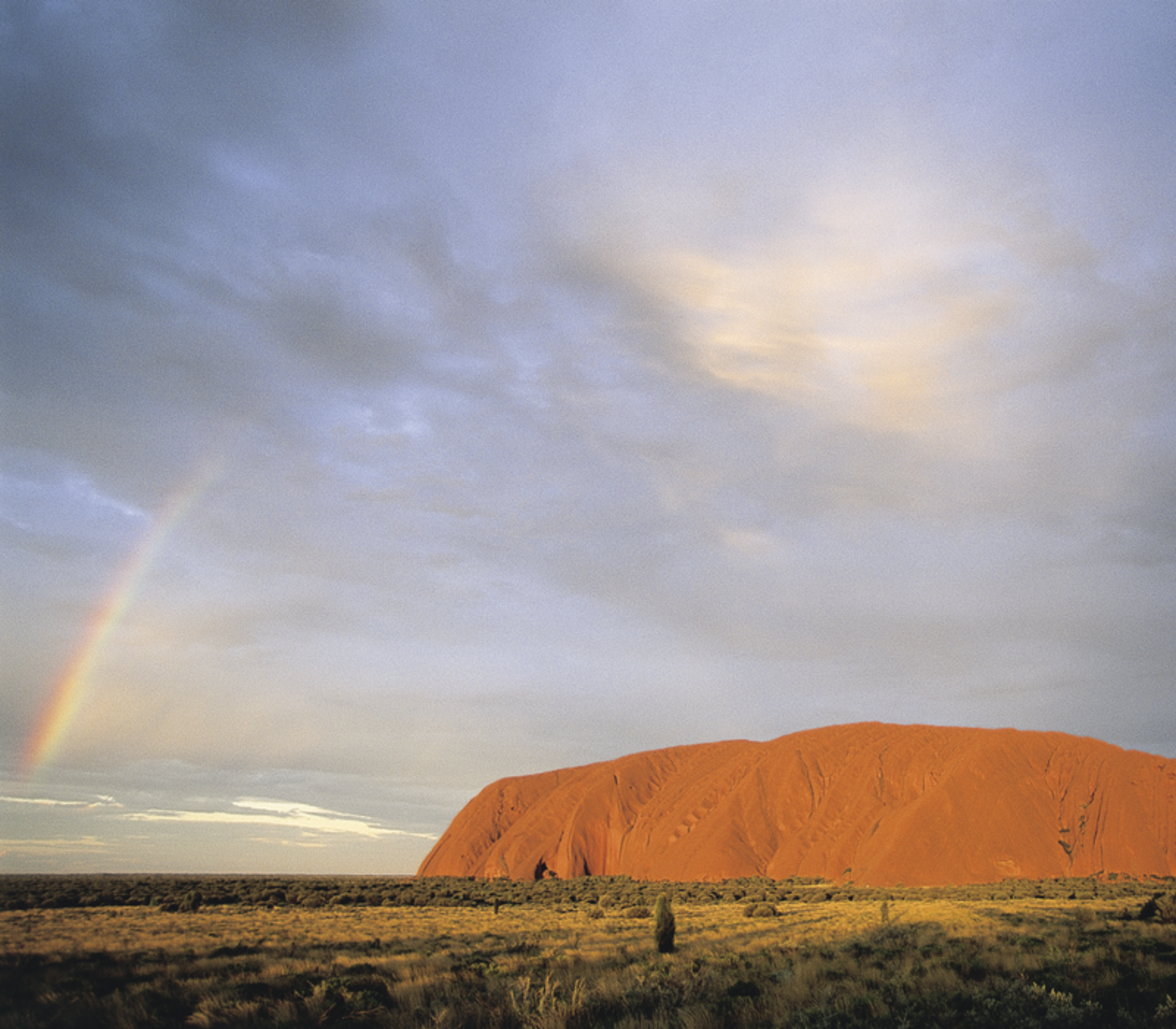 The red monolith of Uluru in the distance with a bright rainbow appearing on the left side under a cloudy sky.