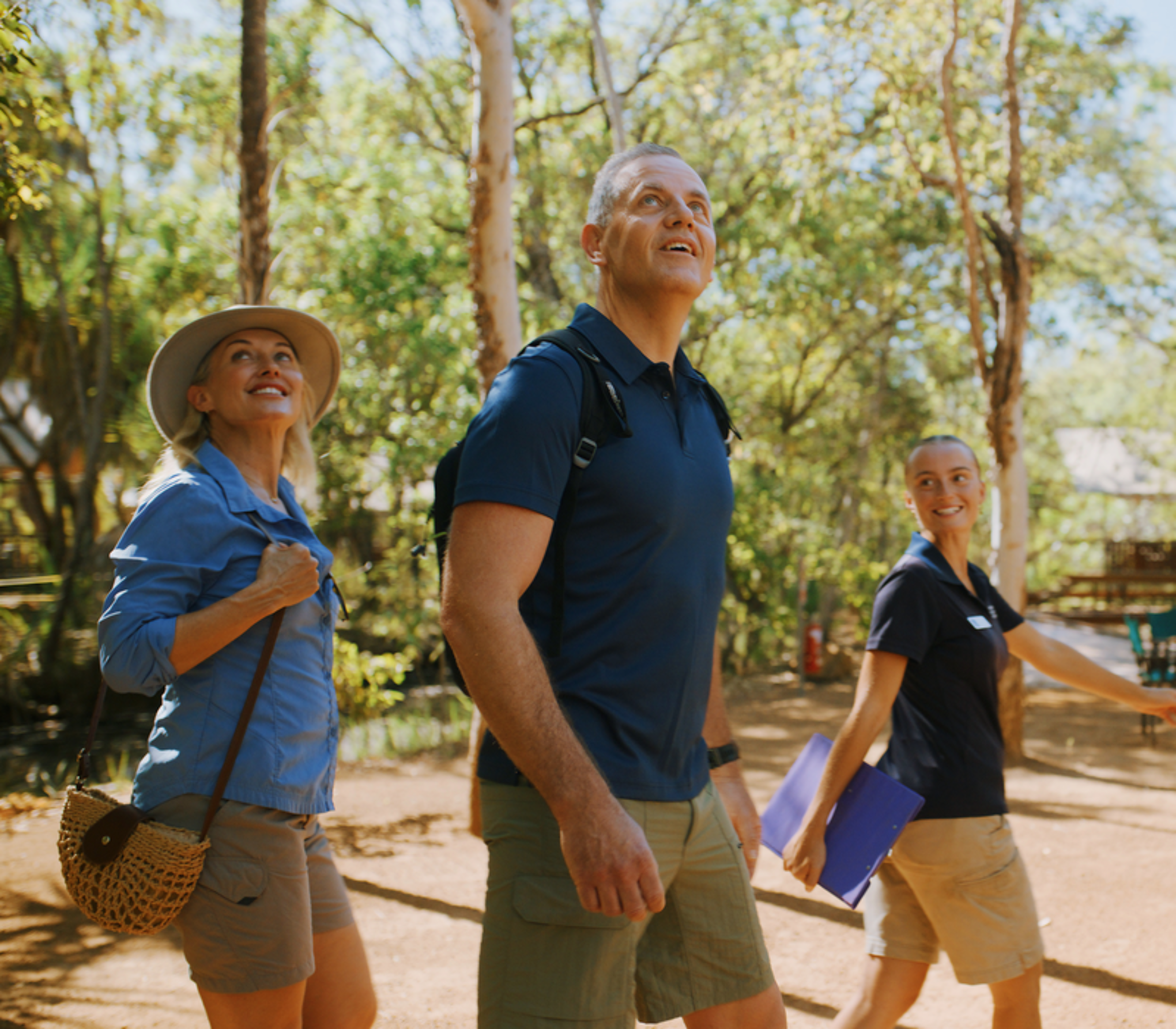 A smiling couple with backpacks walking through a sunlit forest clearing toward a premium glamping tent with a lodge staff member.