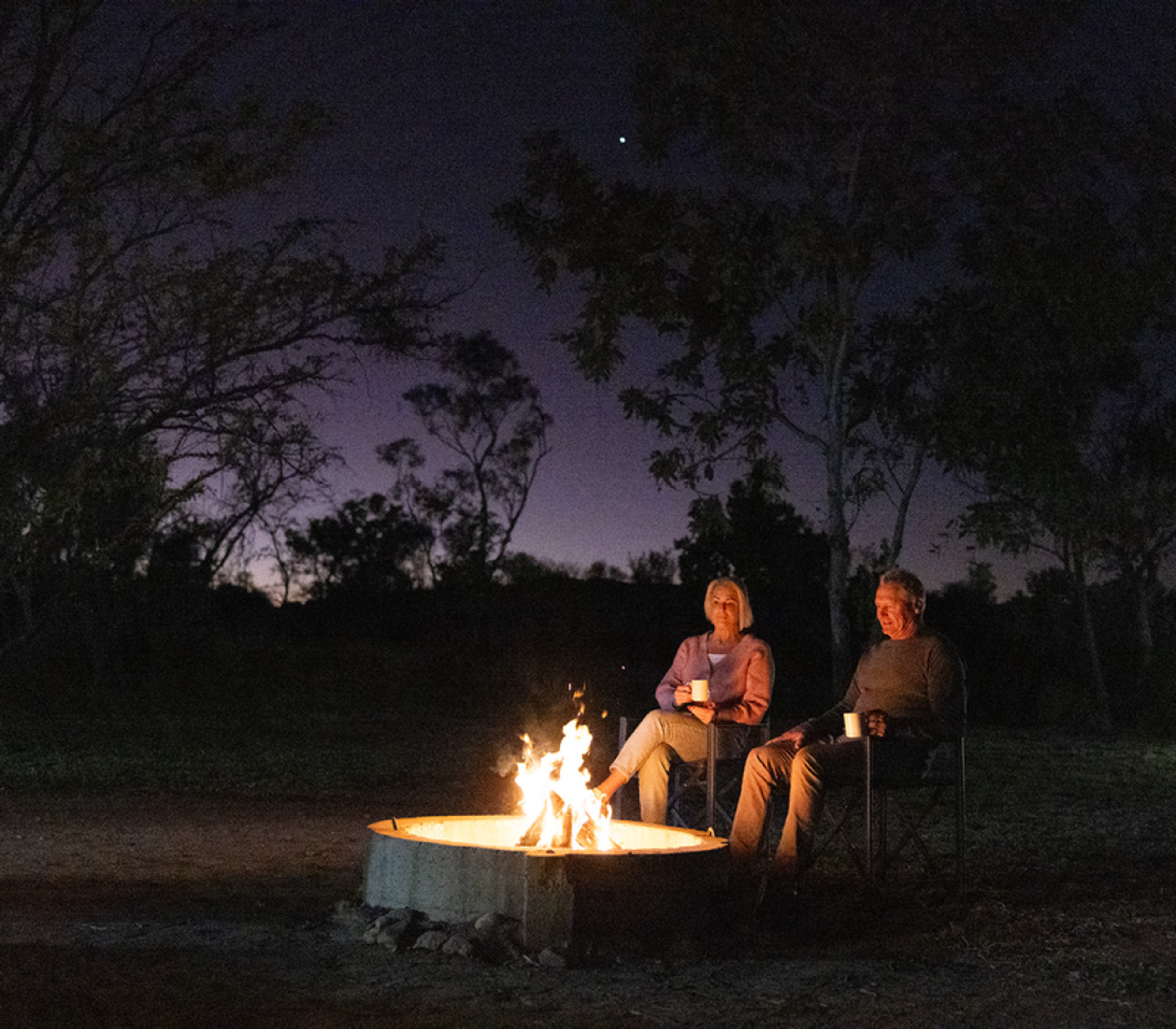 Two people sitting in chairs around a bright campfire at night under a purple dusk sky in the Kimberley.