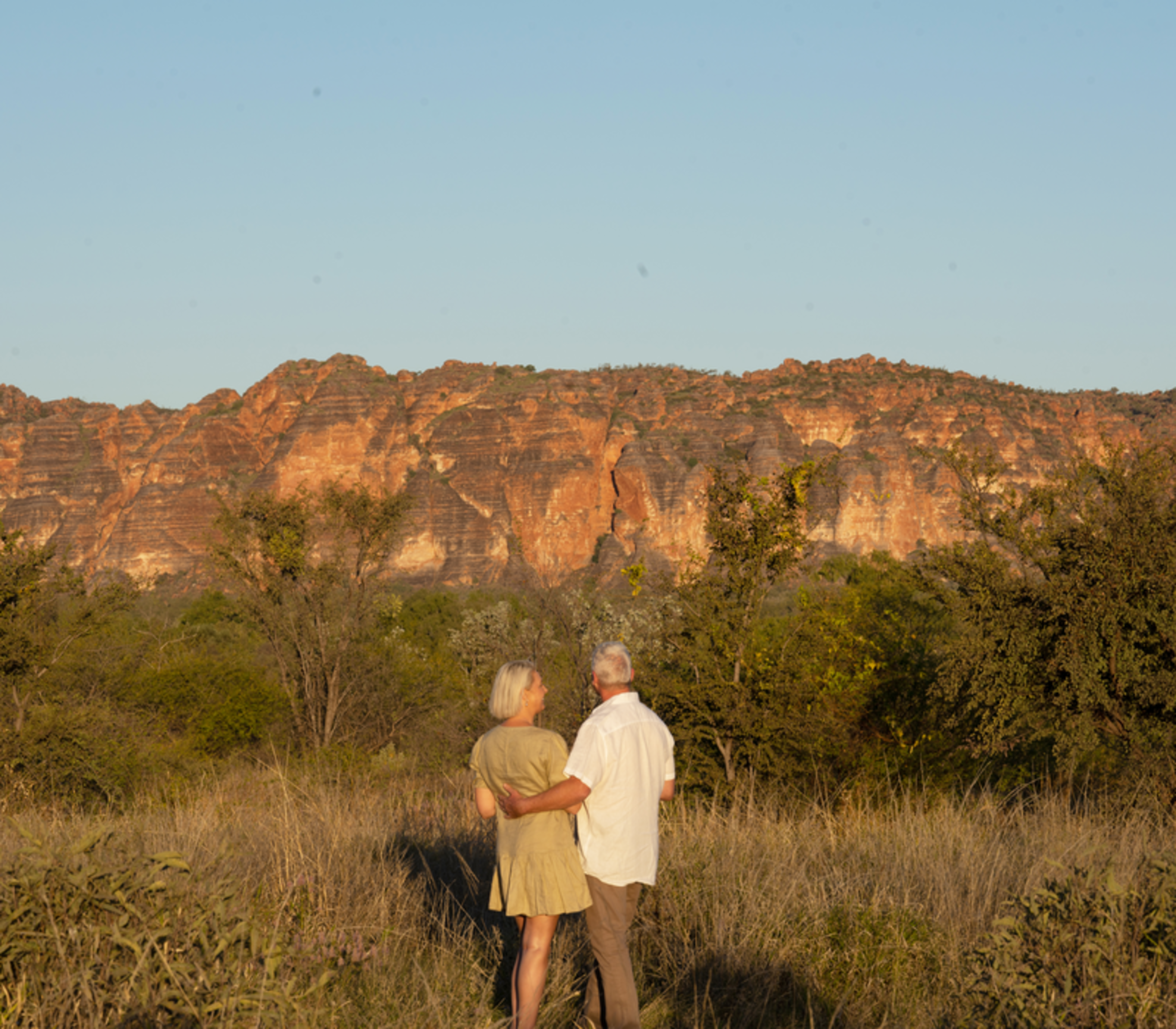 A couple standing in tall grass looking toward the massive striped rock formations of the Bungle Bungle Range.
