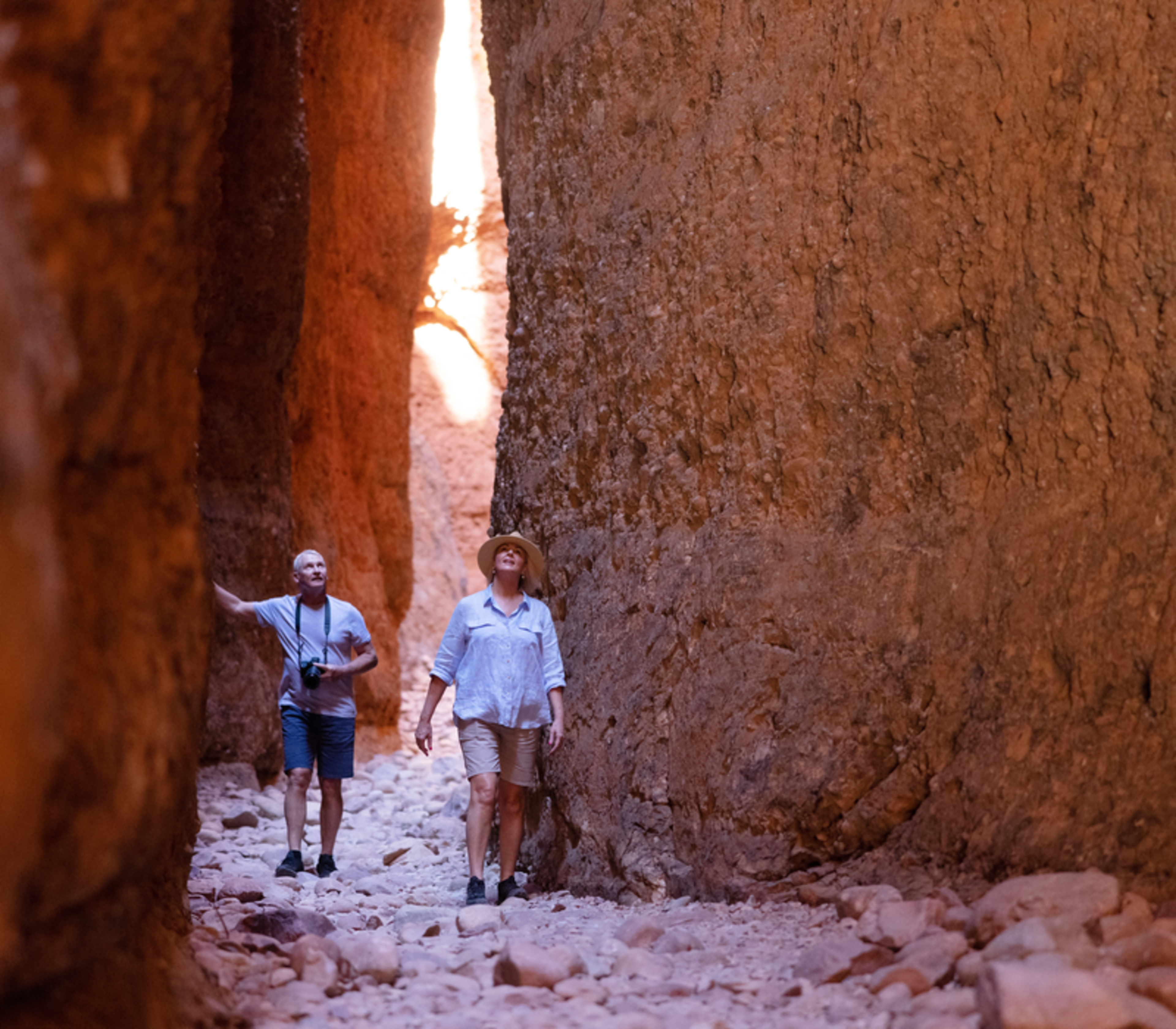 People walking on a rocky path through a very narrow and tall slot canyon with red stone walls.