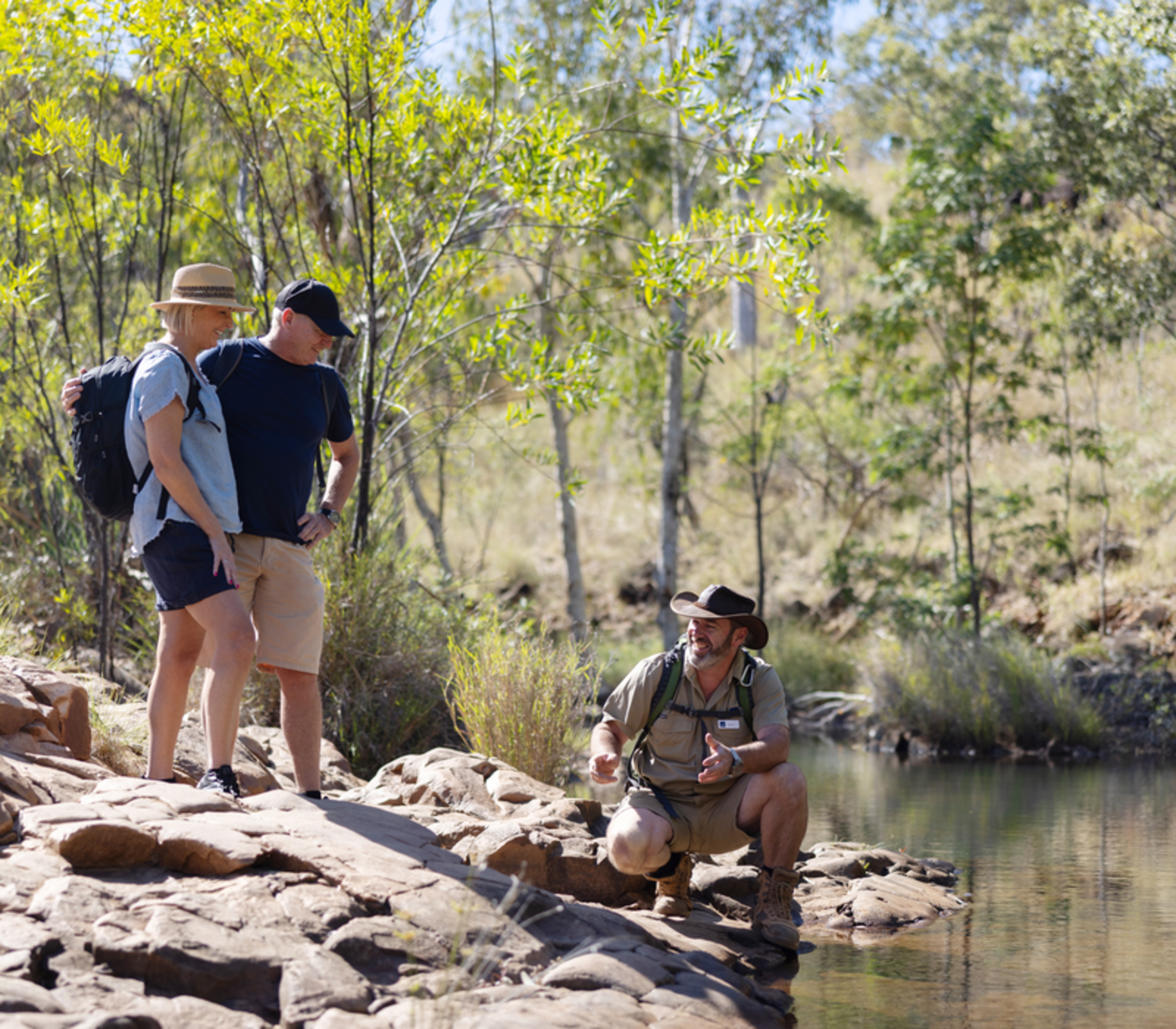 A tour guide in uniform talks to two travelers by a still river in the rocky Kimberley outback.