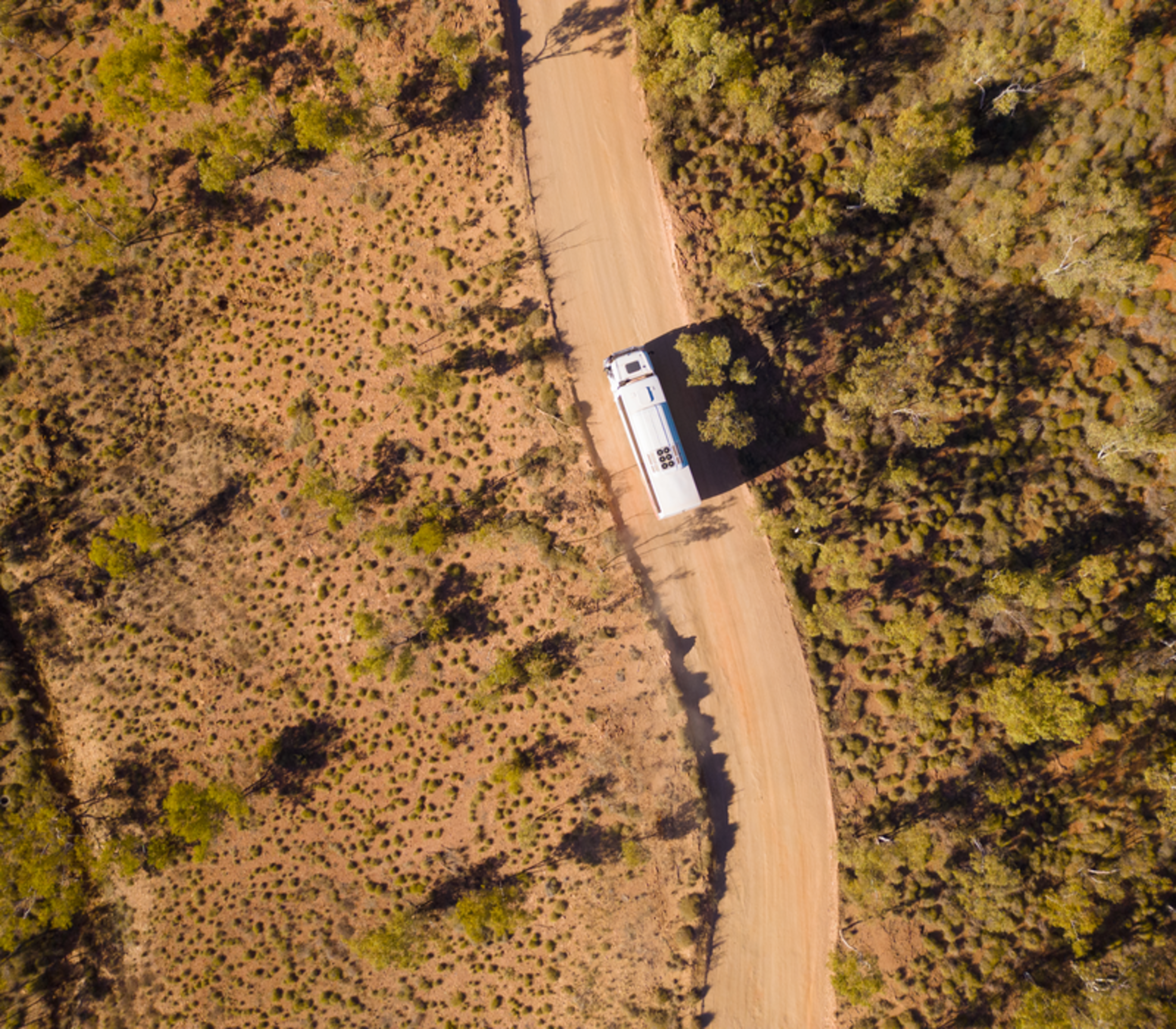Top-down aerial shot of a white tour bus driving on a dusty orange outback road surrounded by green shrubs in Western Australia.