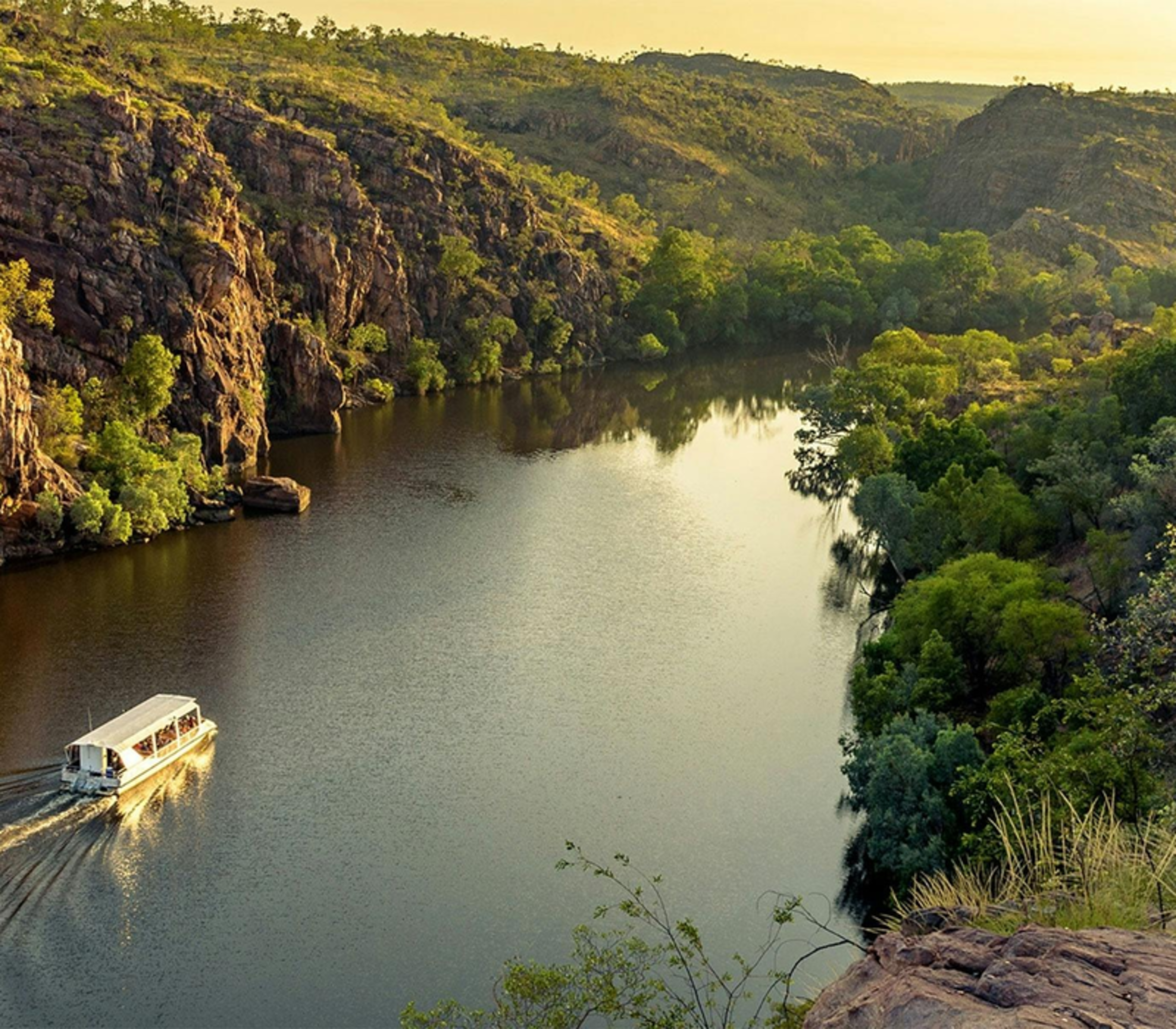 A white tour boat cruising down a river between high rocky cliffs and green trees in Katherine, Northern Territory.