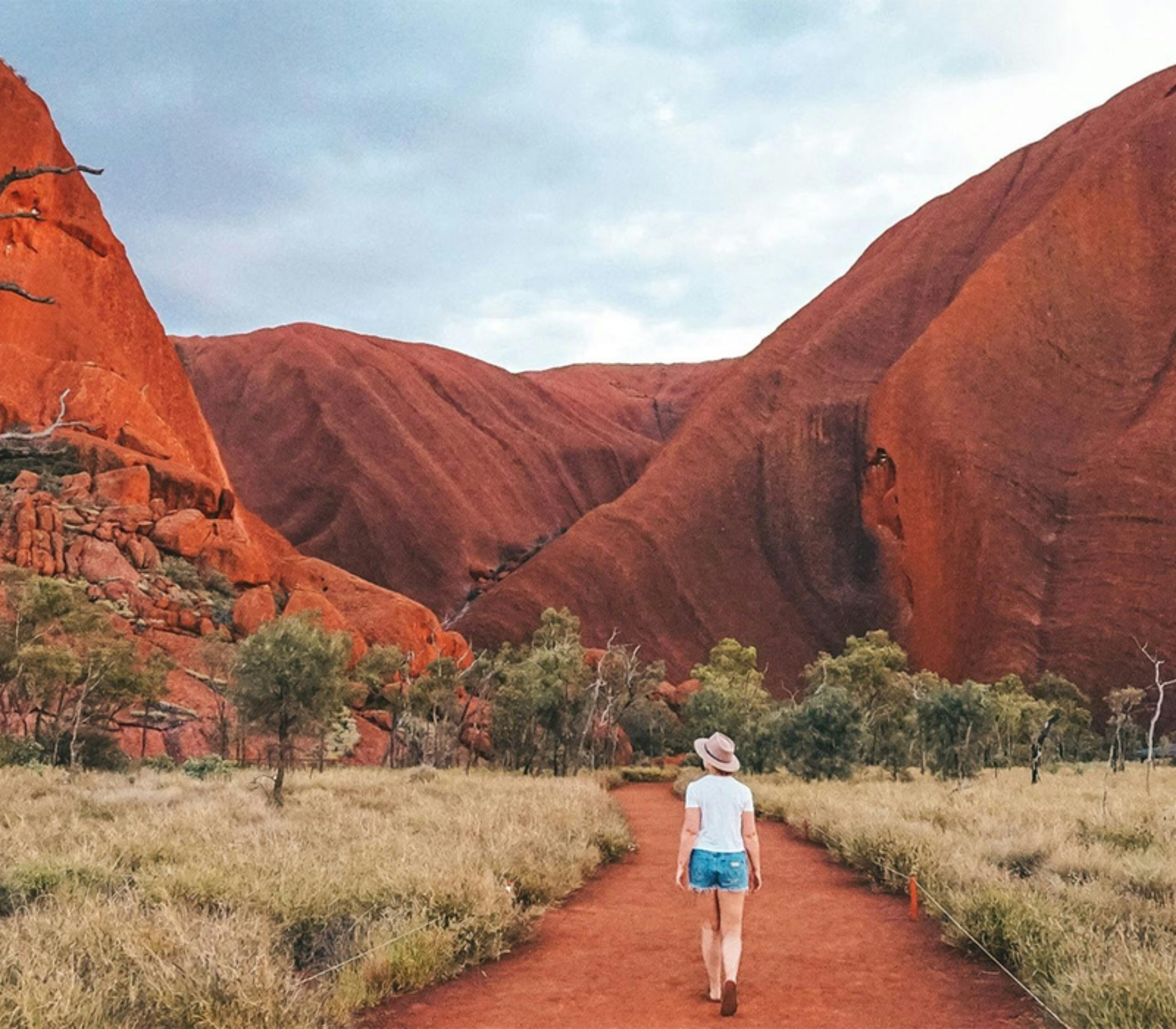 A woman in a hat walking on a red dirt trail between high red rock cliffs under a cloudy sky in the Northern Territory.