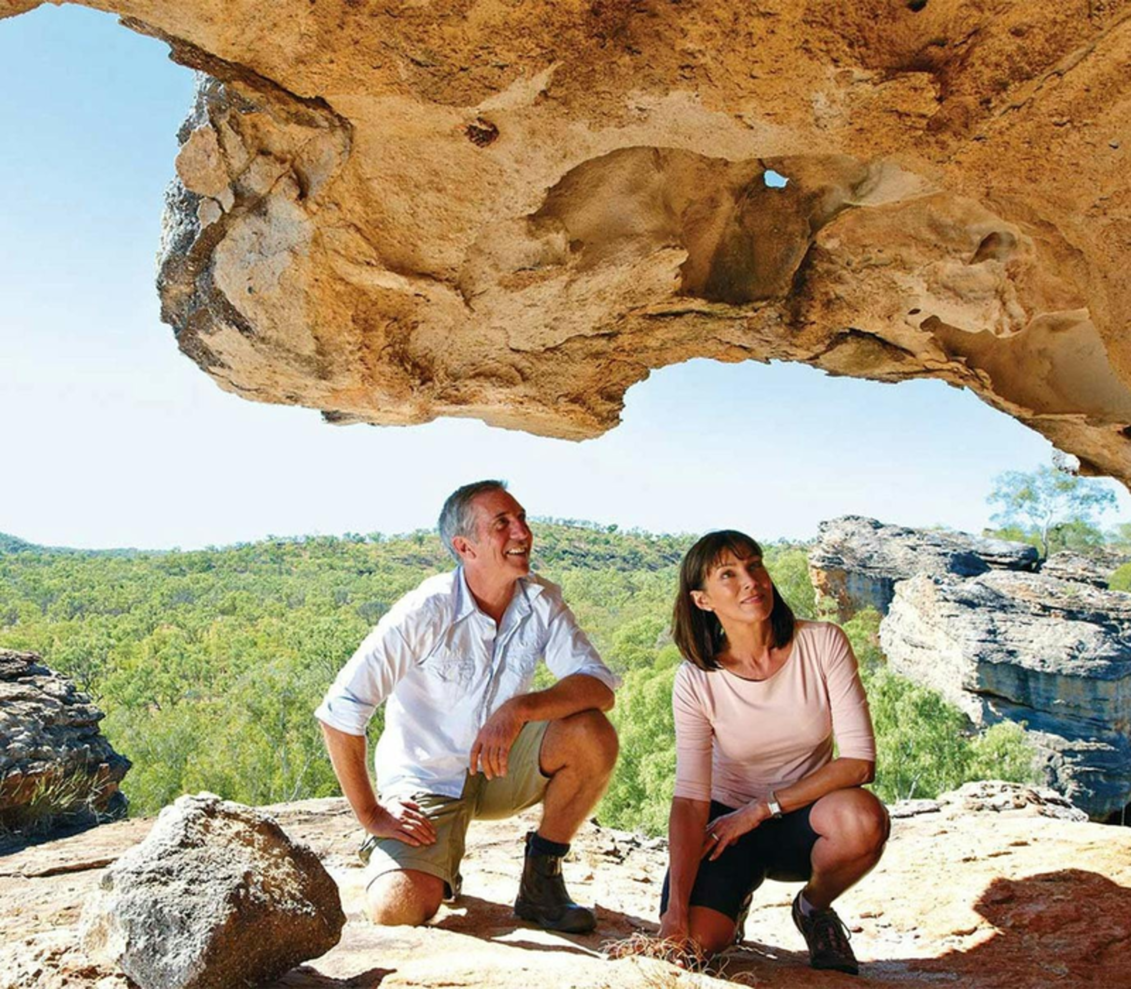 A man and woman kneeling inside a rock cave looking out at the sunlit scrub of the outback.