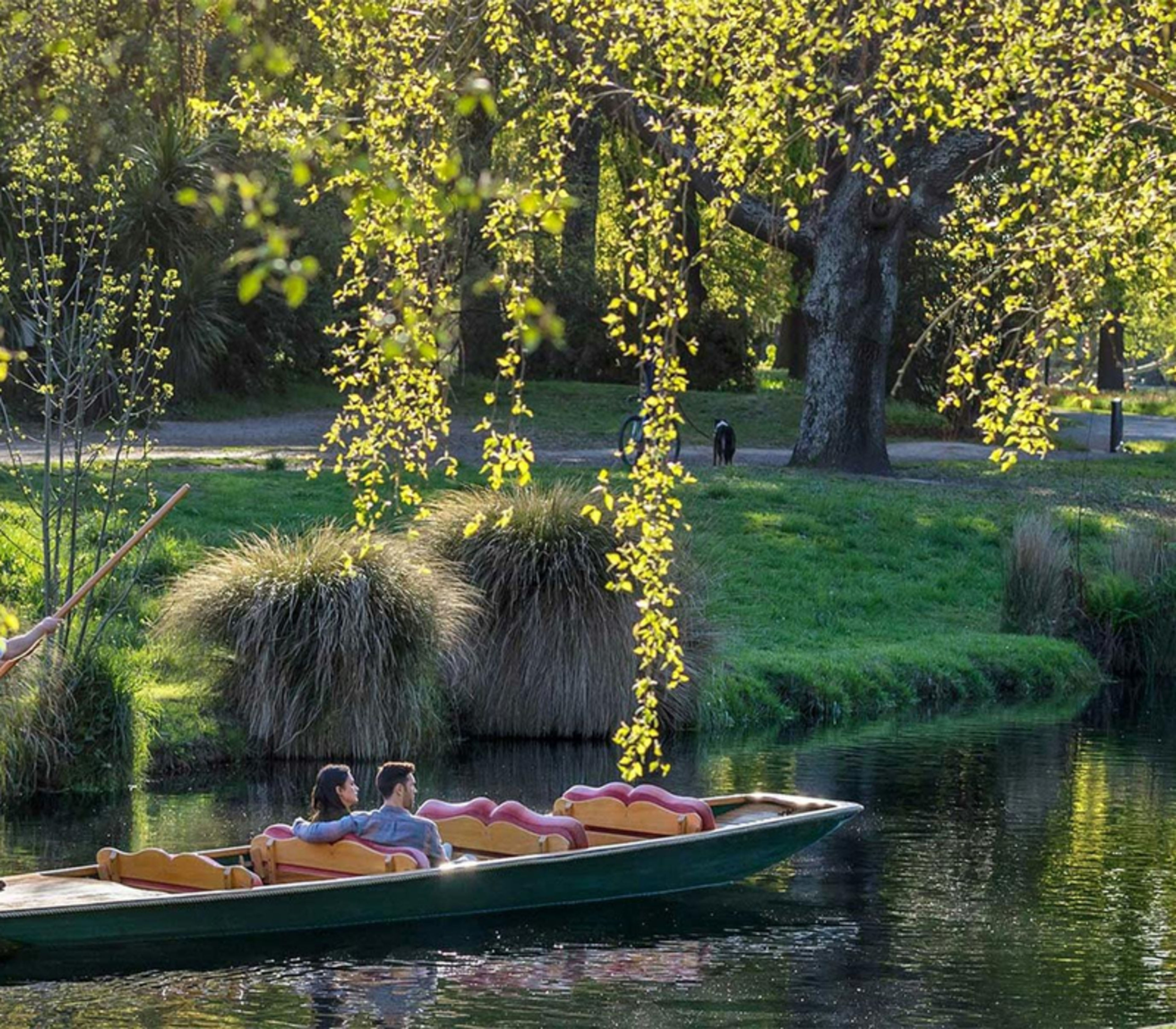 A traditional wooden punt boat with a punter and two passengers gliding down a tree-lined river in a sunlit park.