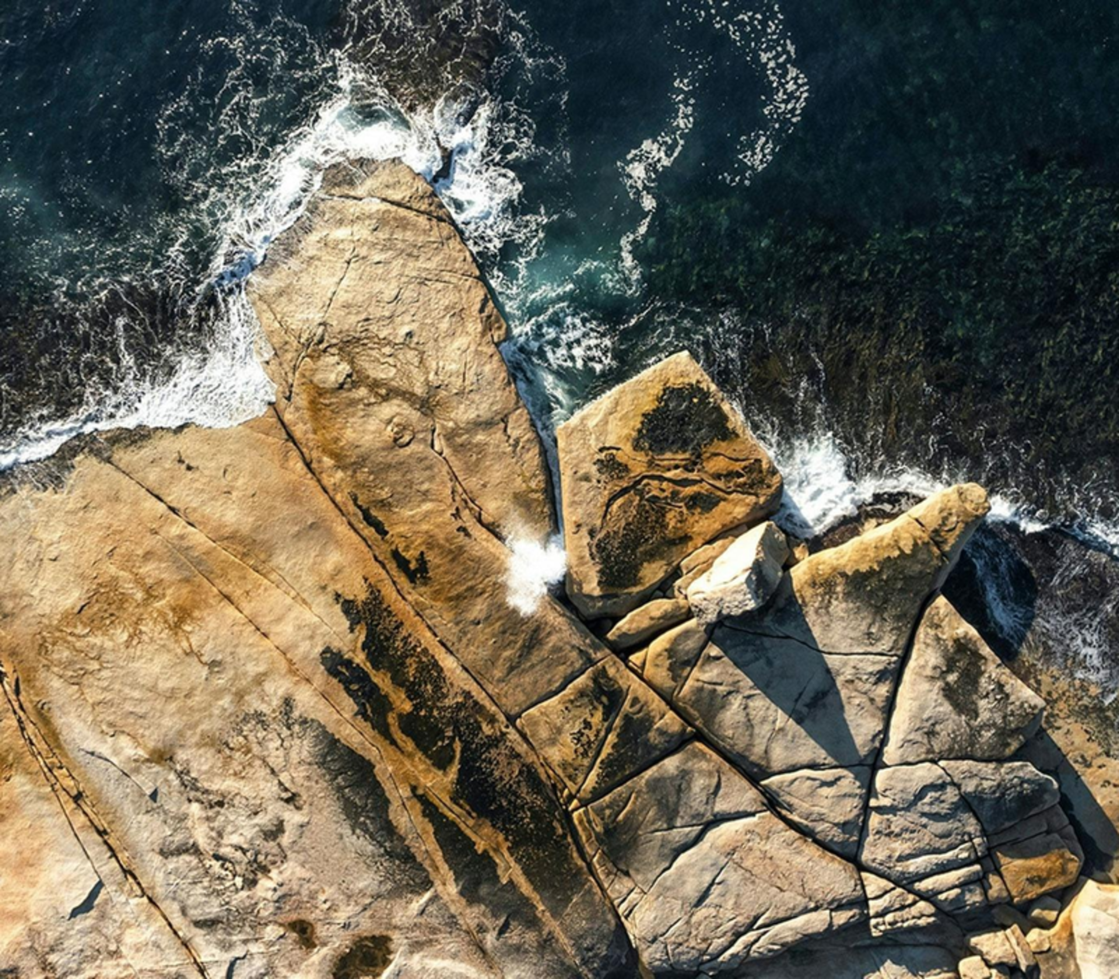 Aerial view looking directly down at waves splashing through a rock formation on a rugged granite shore in Tasmania.