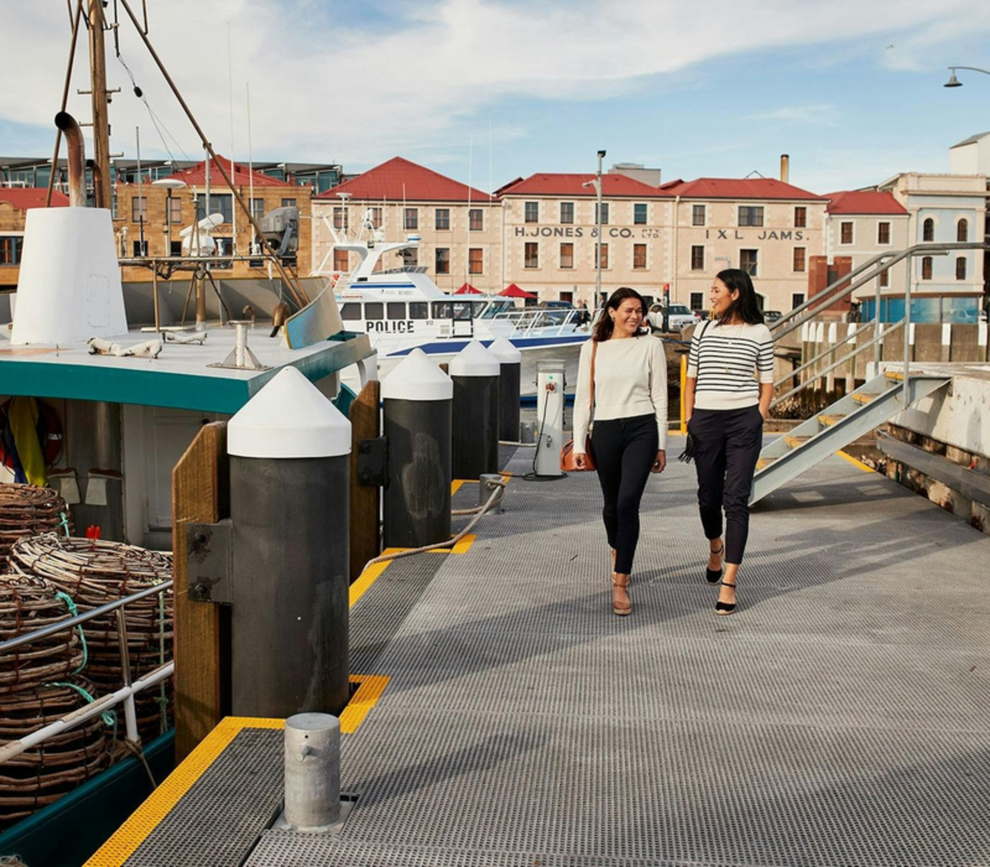 Two women walking on a pier at Hobart harbor with fishing boats and historic stone buildings in the background.