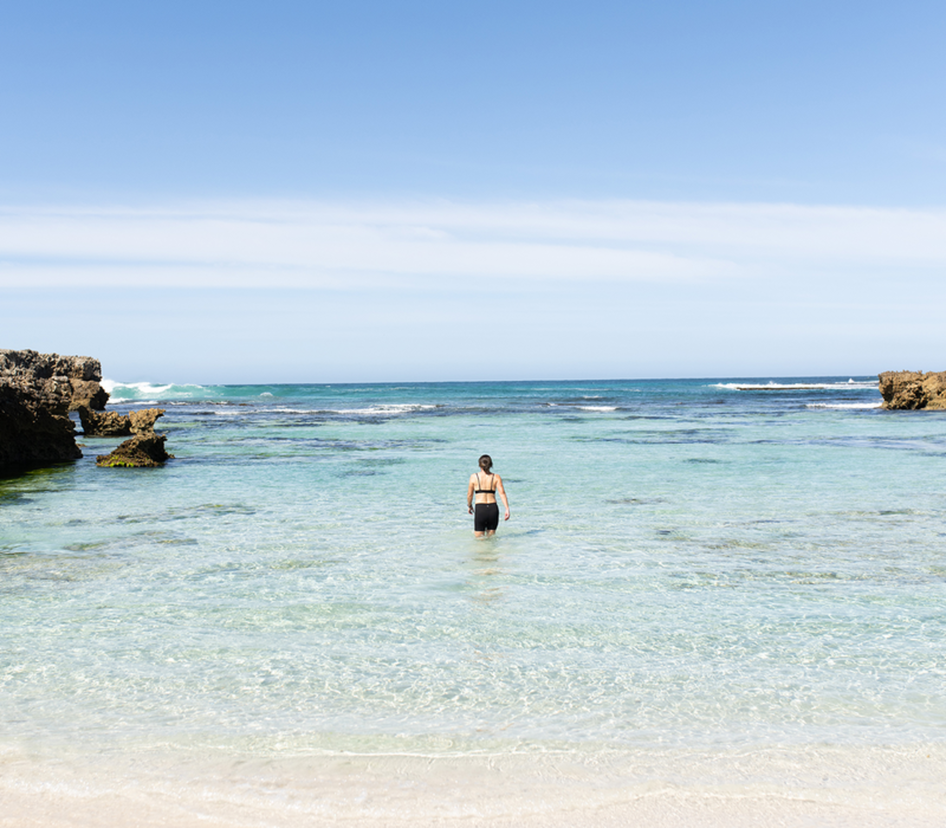 A woman in swimwear wading into clear turquoise ocean water between rocks.