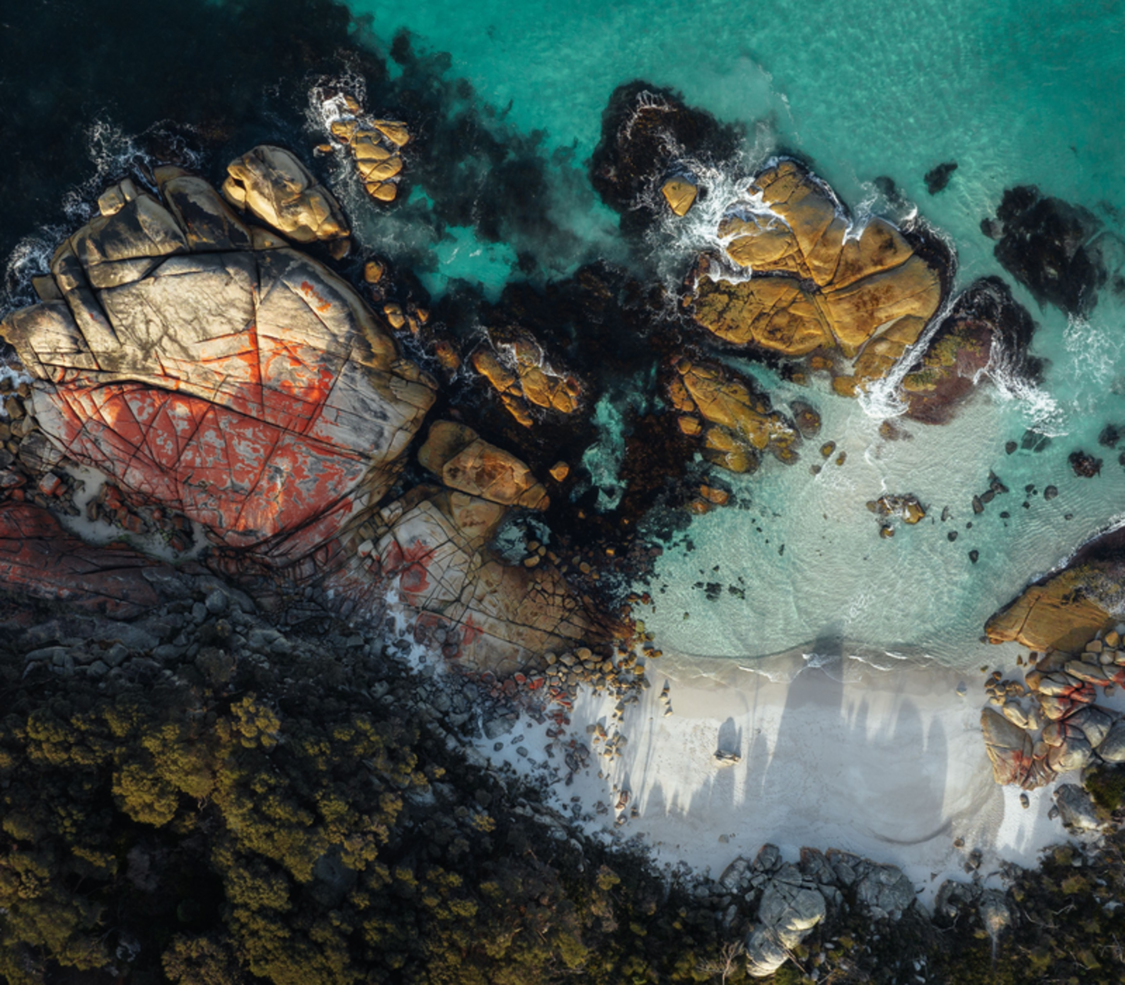 A vertical aerial shot of orange-stained granite boulders meeting crystal clear turquoise ocean water.