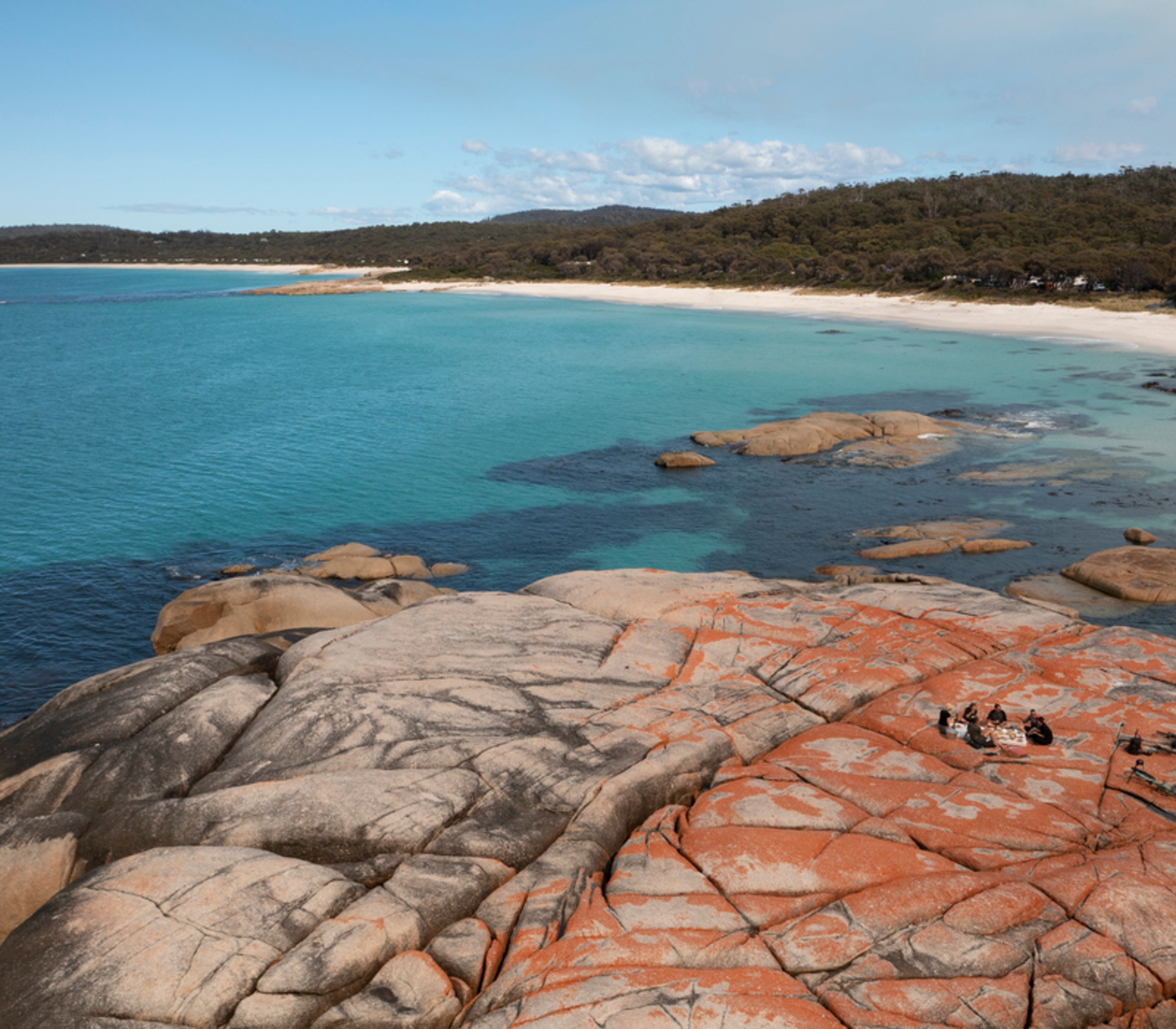 High-angle view of a group sitting on orange-stained rocks next to a white sand beach and turquoise ocean.