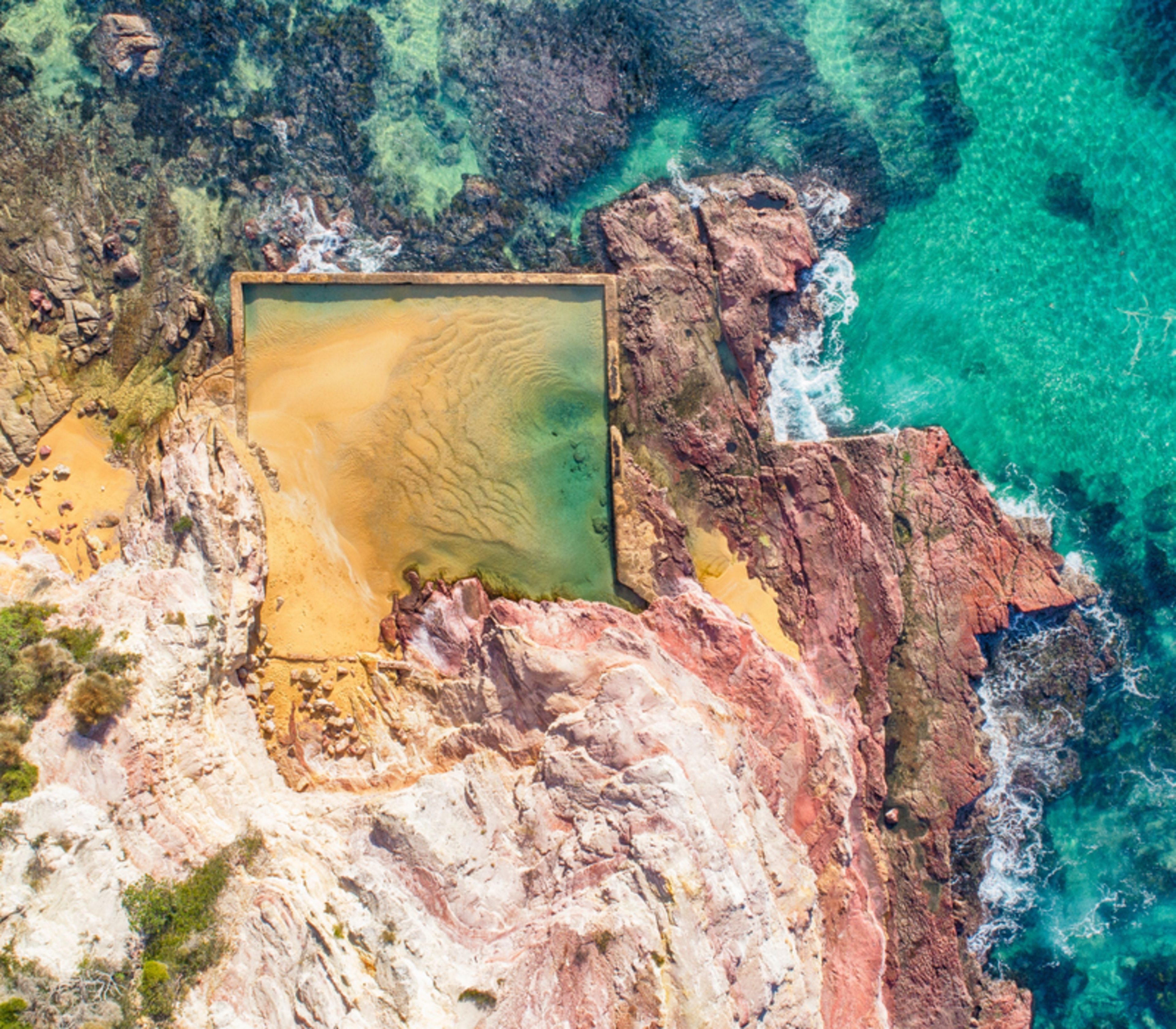 Aerial perspective of a rectangular man-made ocean pool surrounded by rugged red rocks.