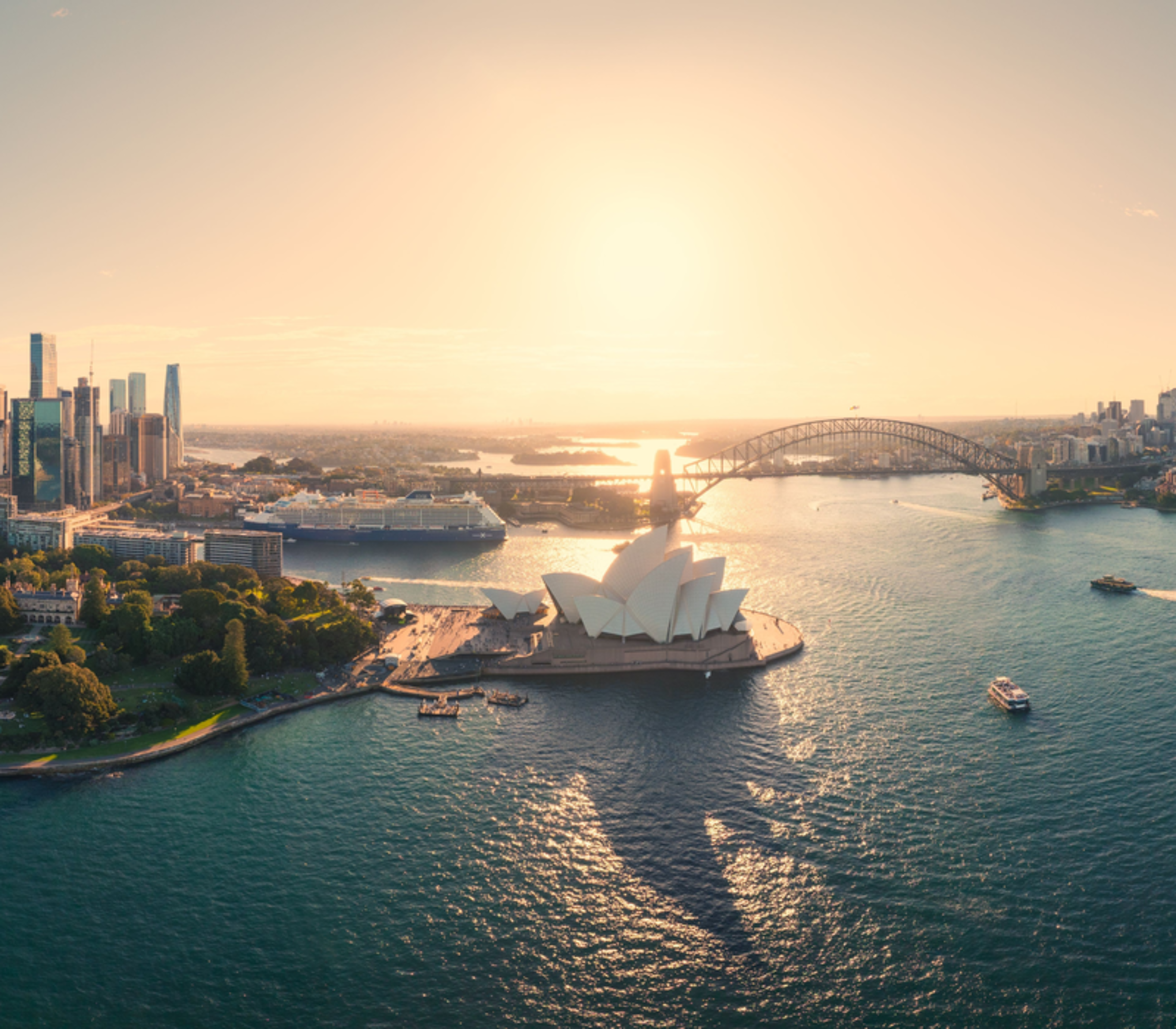Panoramic aerial view of Sydney Harbour, the city skyline, and the Opera House under a golden sun.