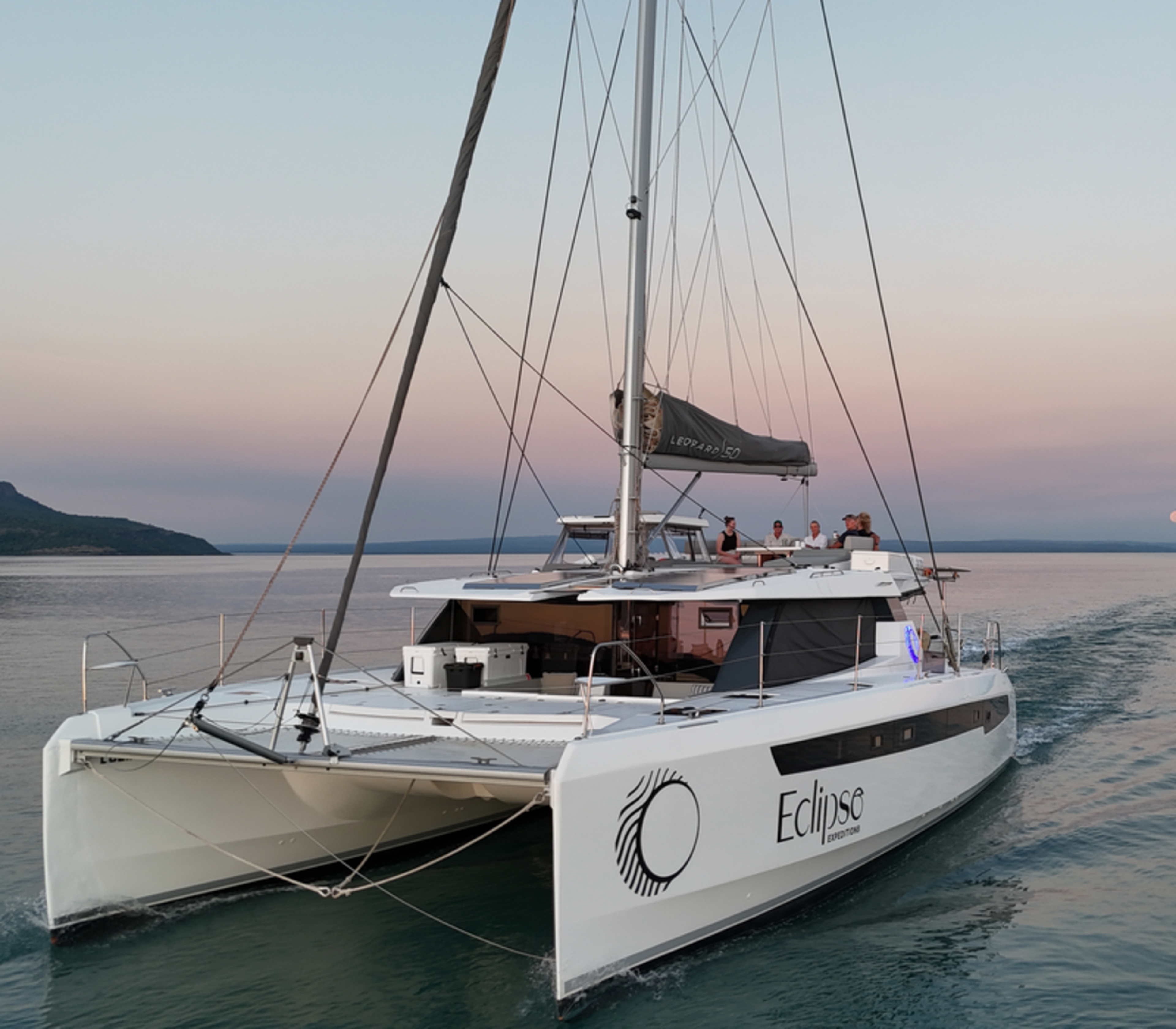 A low-angle shot from the water looking up at the back of the white catamaran Eclipse. Guests are visible on the top deck, with a full moon rising on the horizon under a soft pink and purple sky.