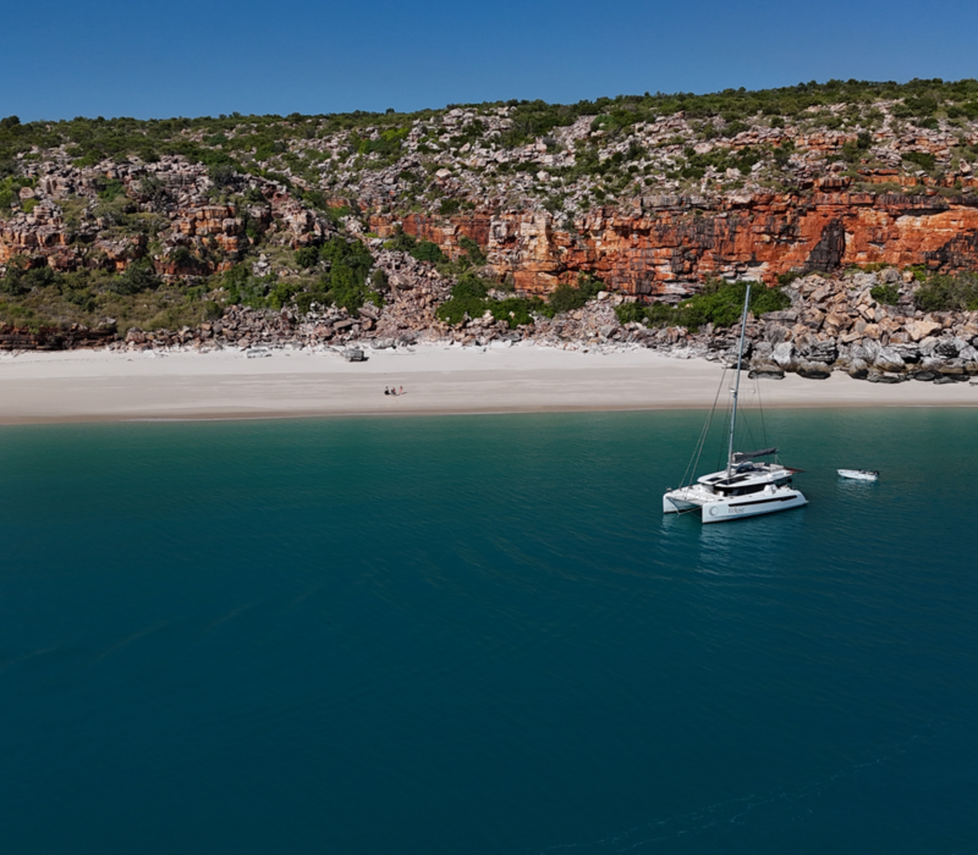 A wide aerial shot of a pristine white sand beach at the base of a tall, layered orange rock cliff. The Eclipse catamaran is anchored in the clear turquoise water just offshore.