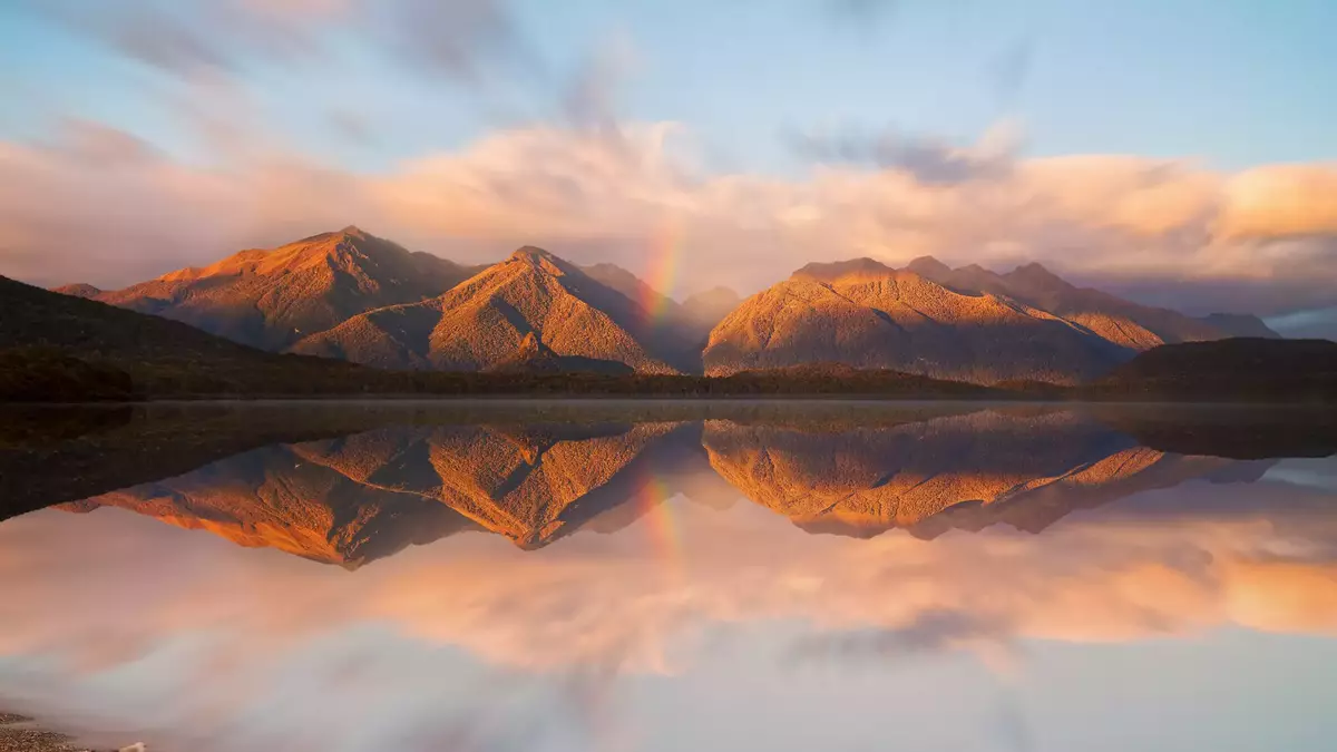 Lake Manapouri reflecting mountains, clouds, and a rainbow at sunset Main Image