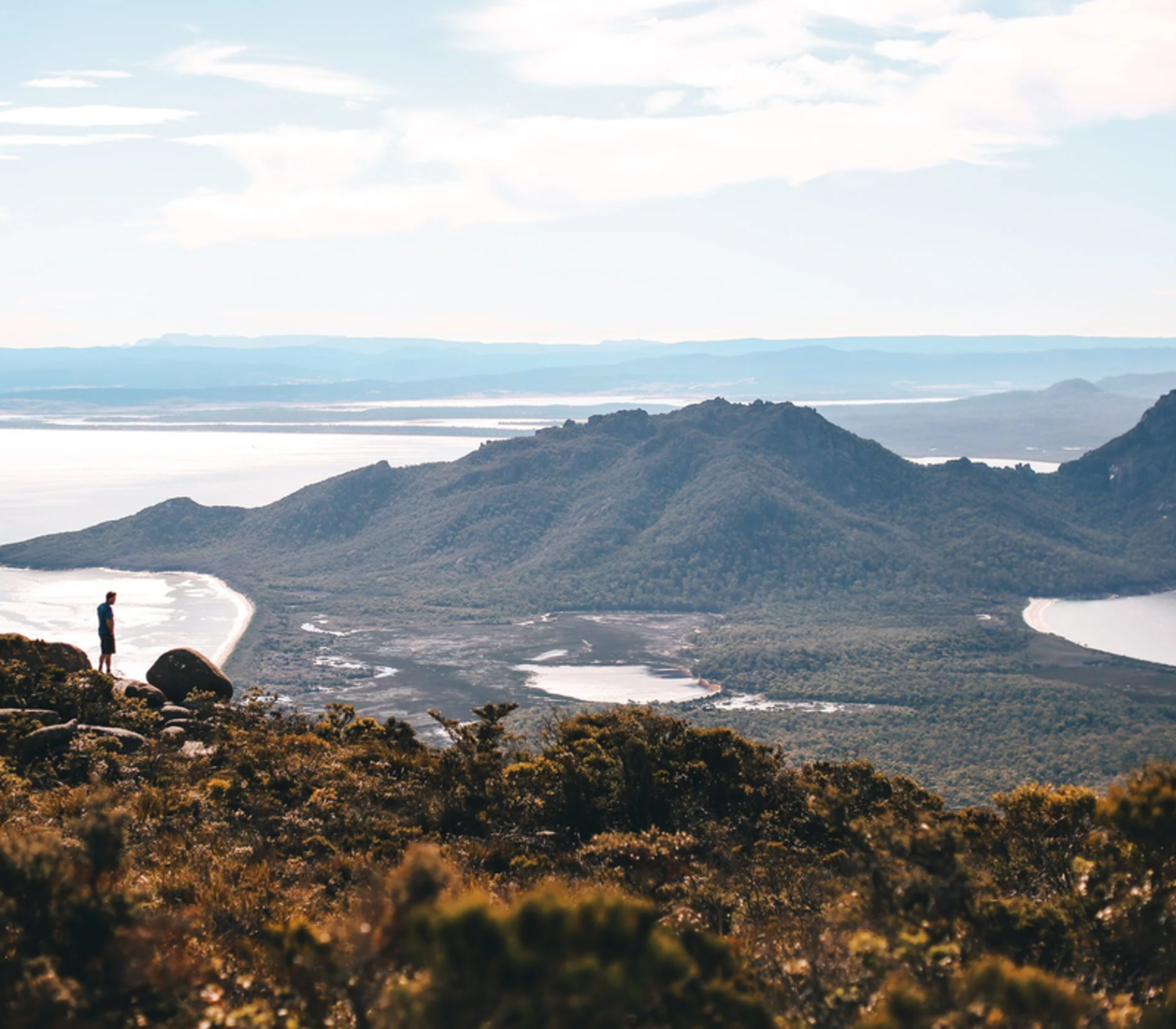 Silhouette of a person standing on a rocky cliff overlooking a coastal landscape with lagoons and mountains