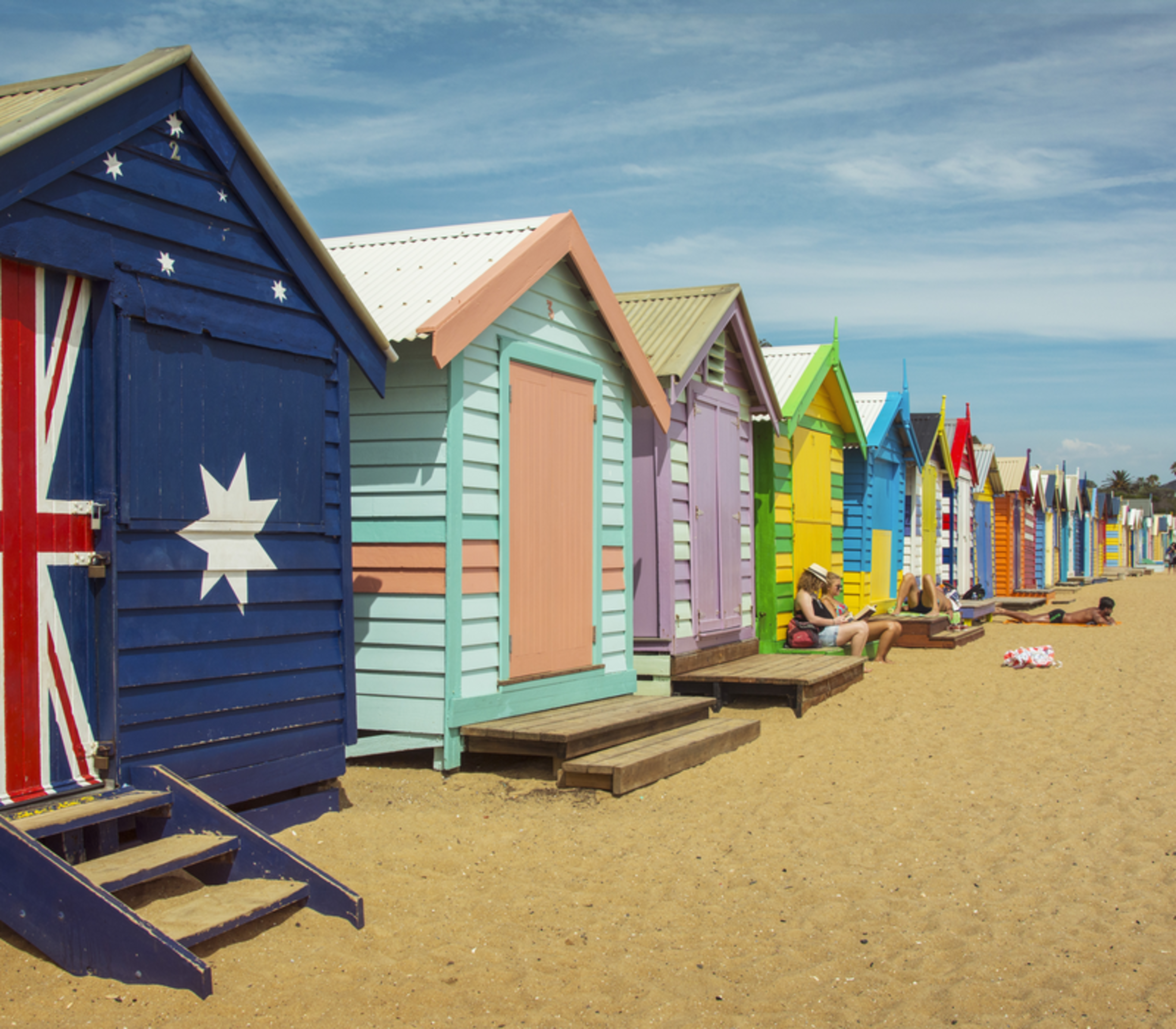 A row of brightly painted wooden beach huts on the sand under a blue sky in Melbourne.