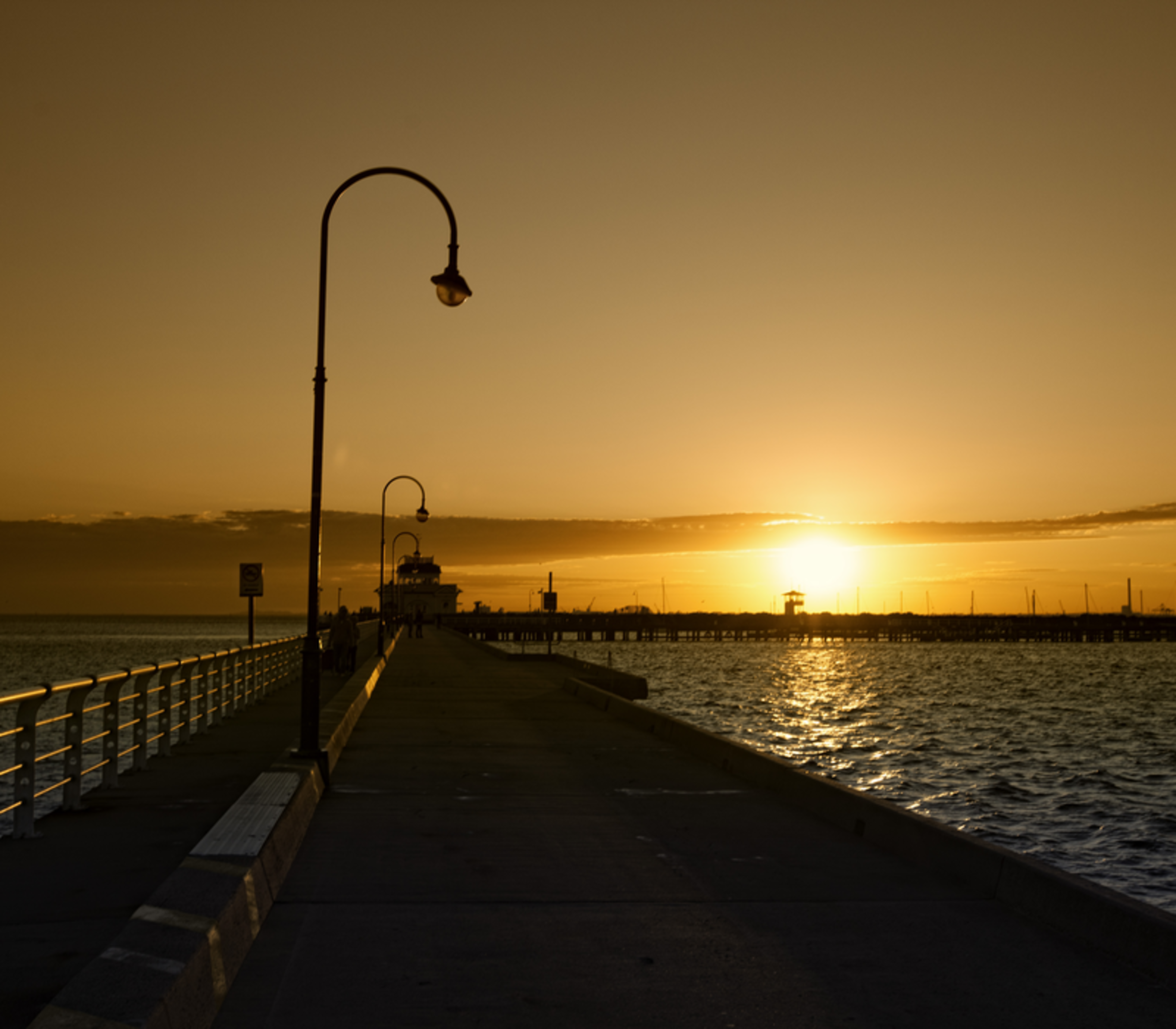 Silhouetted street lamps on a concrete pier leading toward a vibrant orange sunset over the ocean in Melbourne.