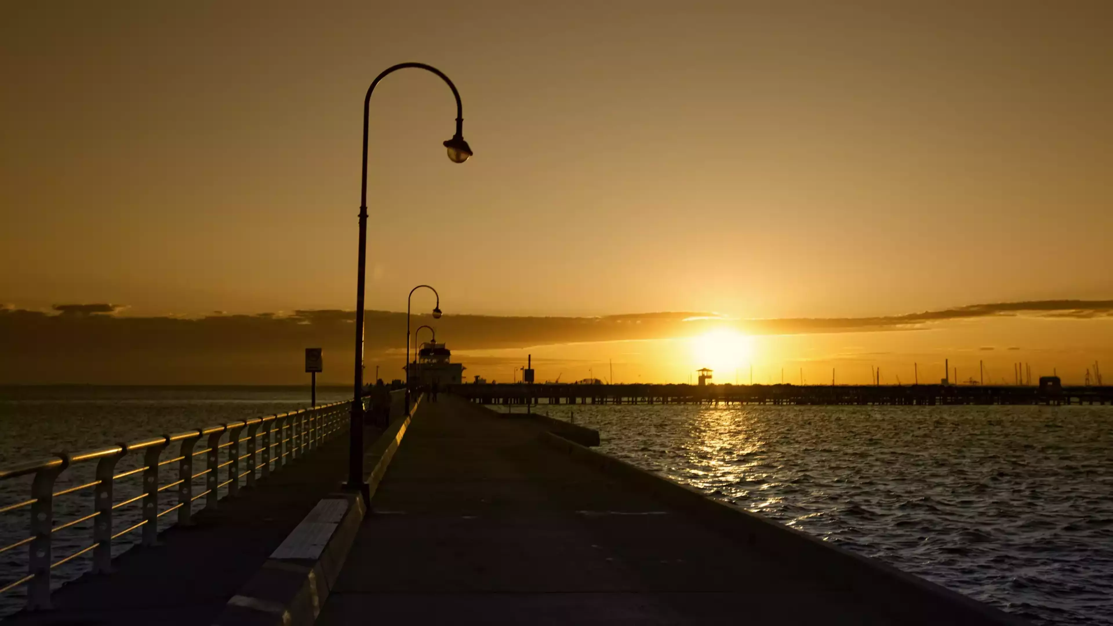 Silhouetted street lamps on a concrete pier leading toward a vibrant orange sunset over the ocean in Melbourne.